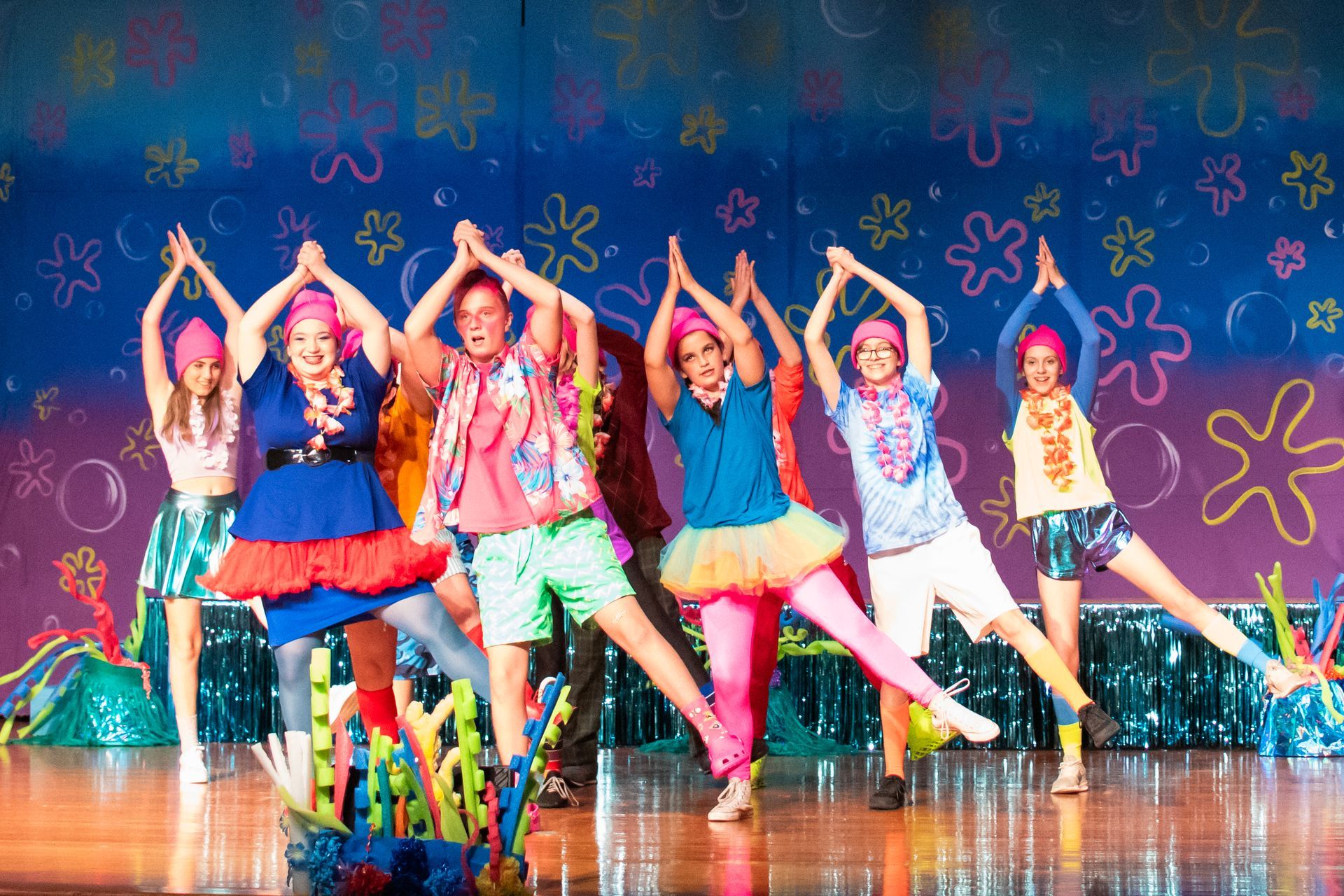 Group of dancers in colorful costumes with arms raised on a stage with a SpongeBob backdrop.