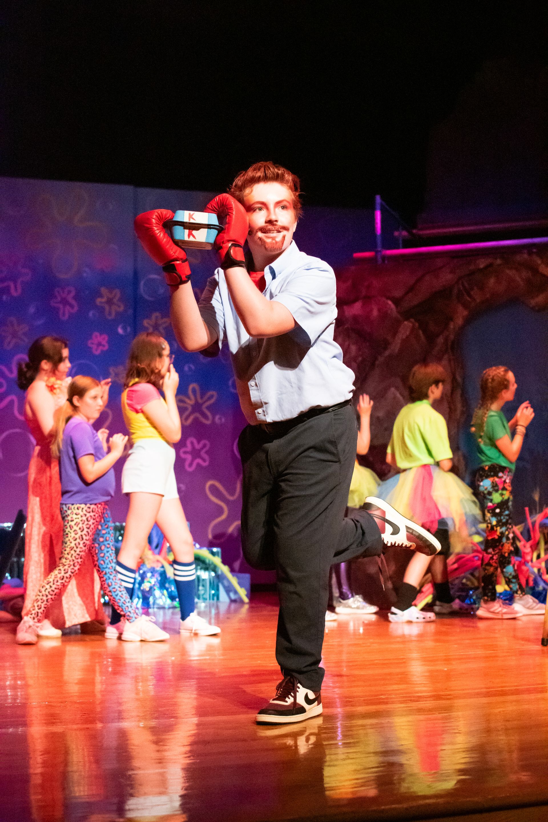 Actor in Mr. Krabs costume holds a box, leg raised on stage with child performers.