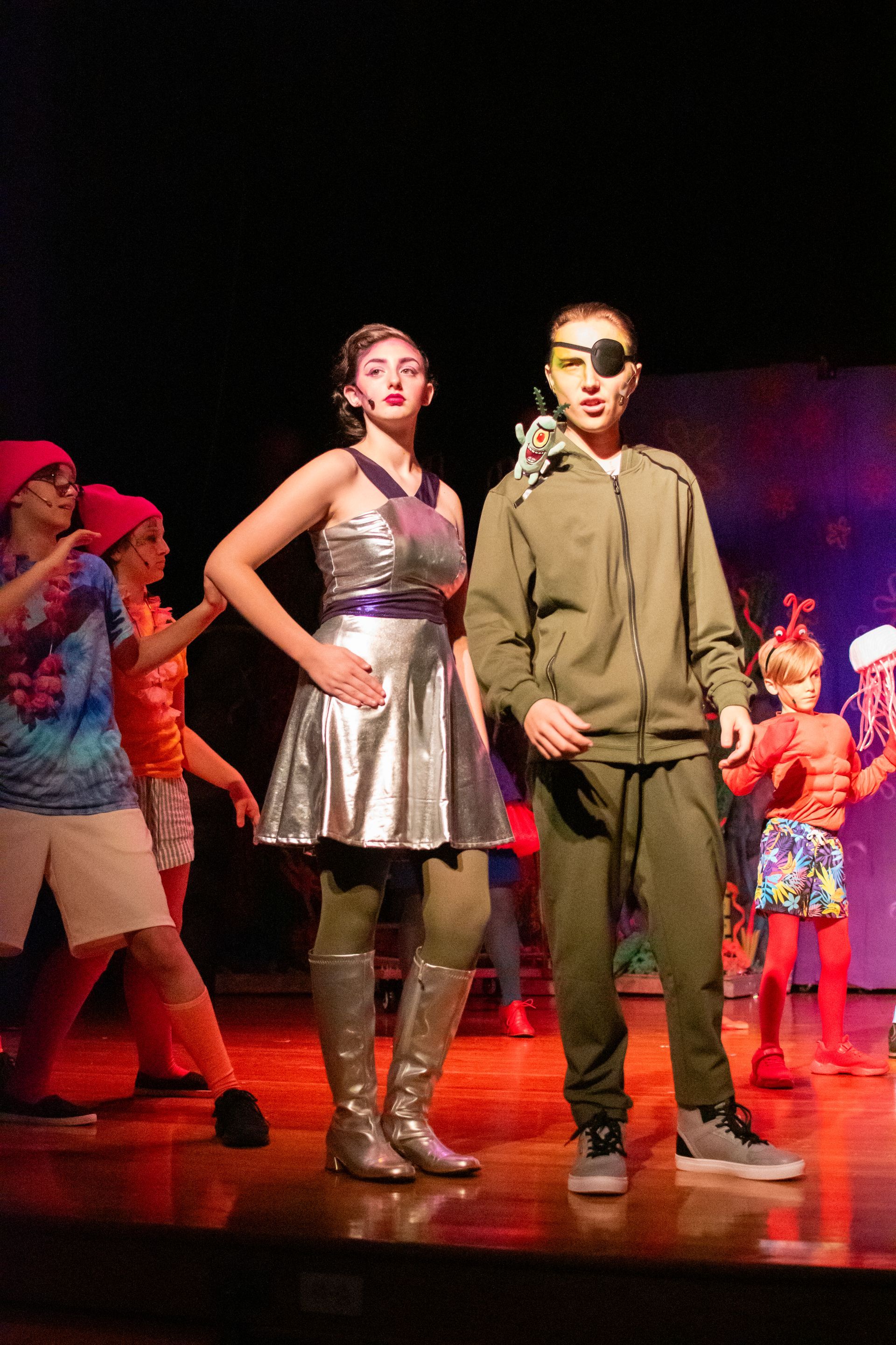 Two people on stage in costume, silver dress and green outfit, eye patch, other performers in background.