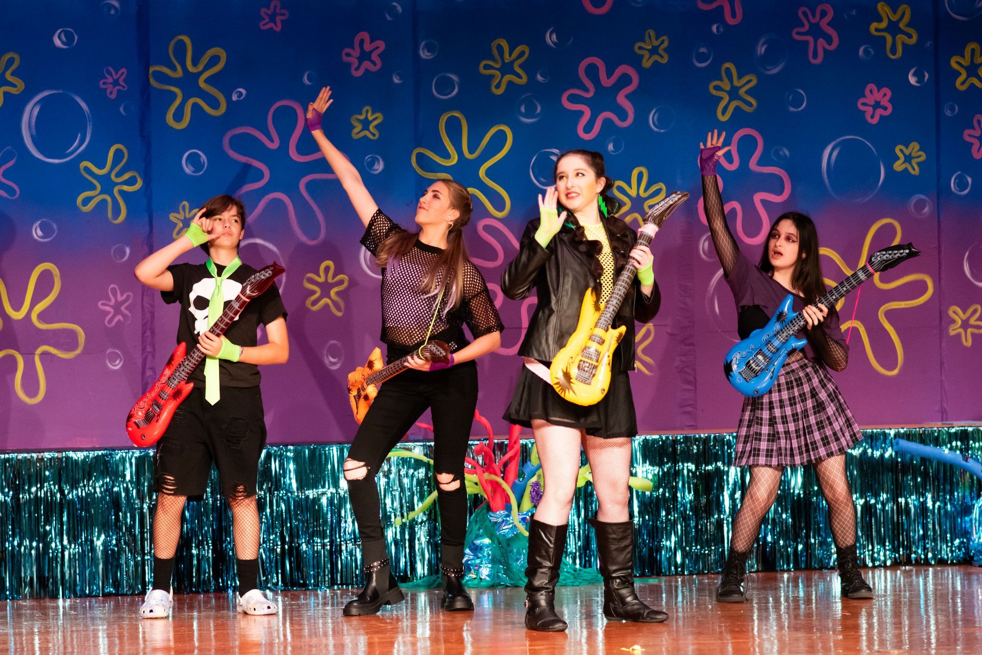 Four young women playing electric guitars on stage, colorful background with splatter and bubbles.