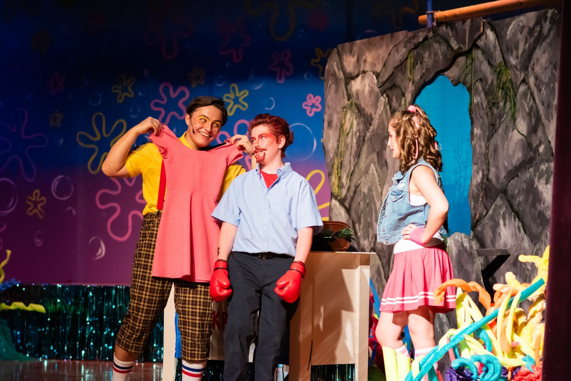 Actors in a stage production of Spongebob Squarepants. One holds up a pink dress, two others look on.