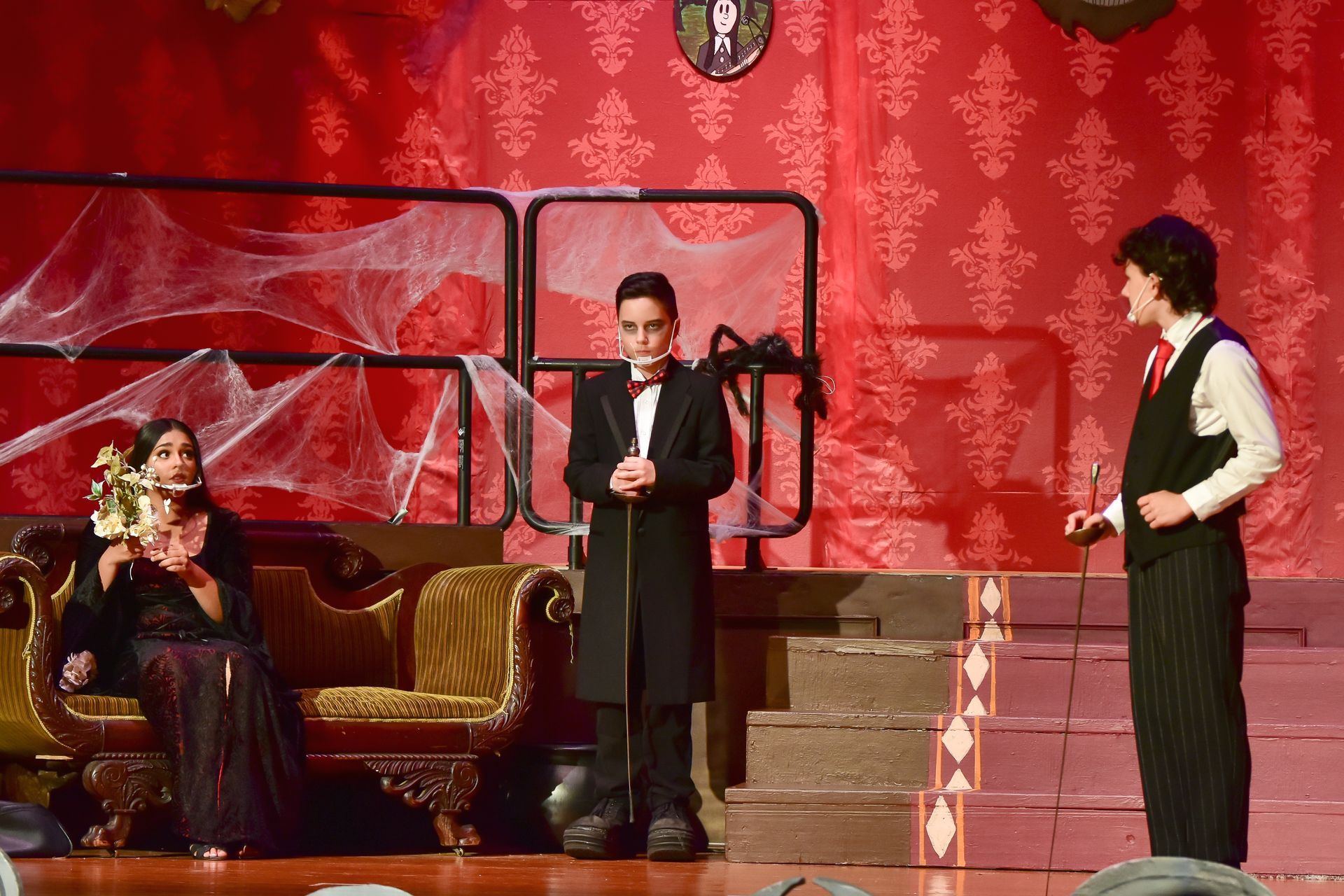 Theater scene: Three actors on stage with gothic backdrop. One holds a microphone.