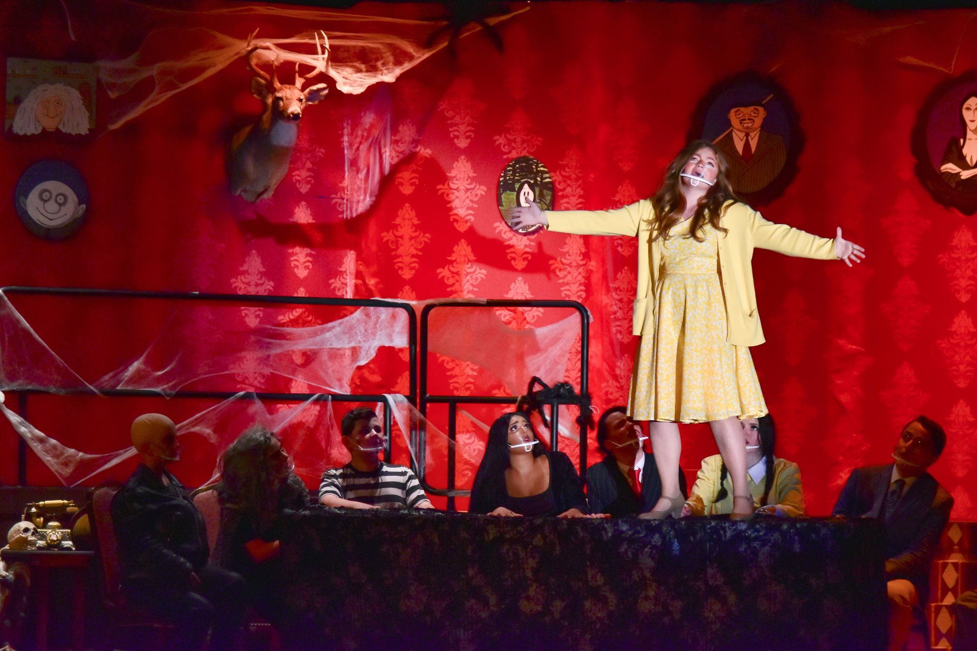 A woman in yellow on stage, arms outstretched, with seated cast. Red backdrop, spooky decor.