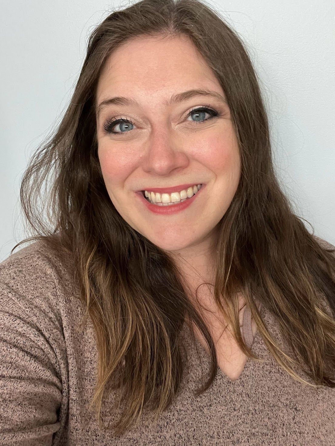 Woman with long brown hair smiles, wearing a pinkish-brown sweater, looking directly at the camera.