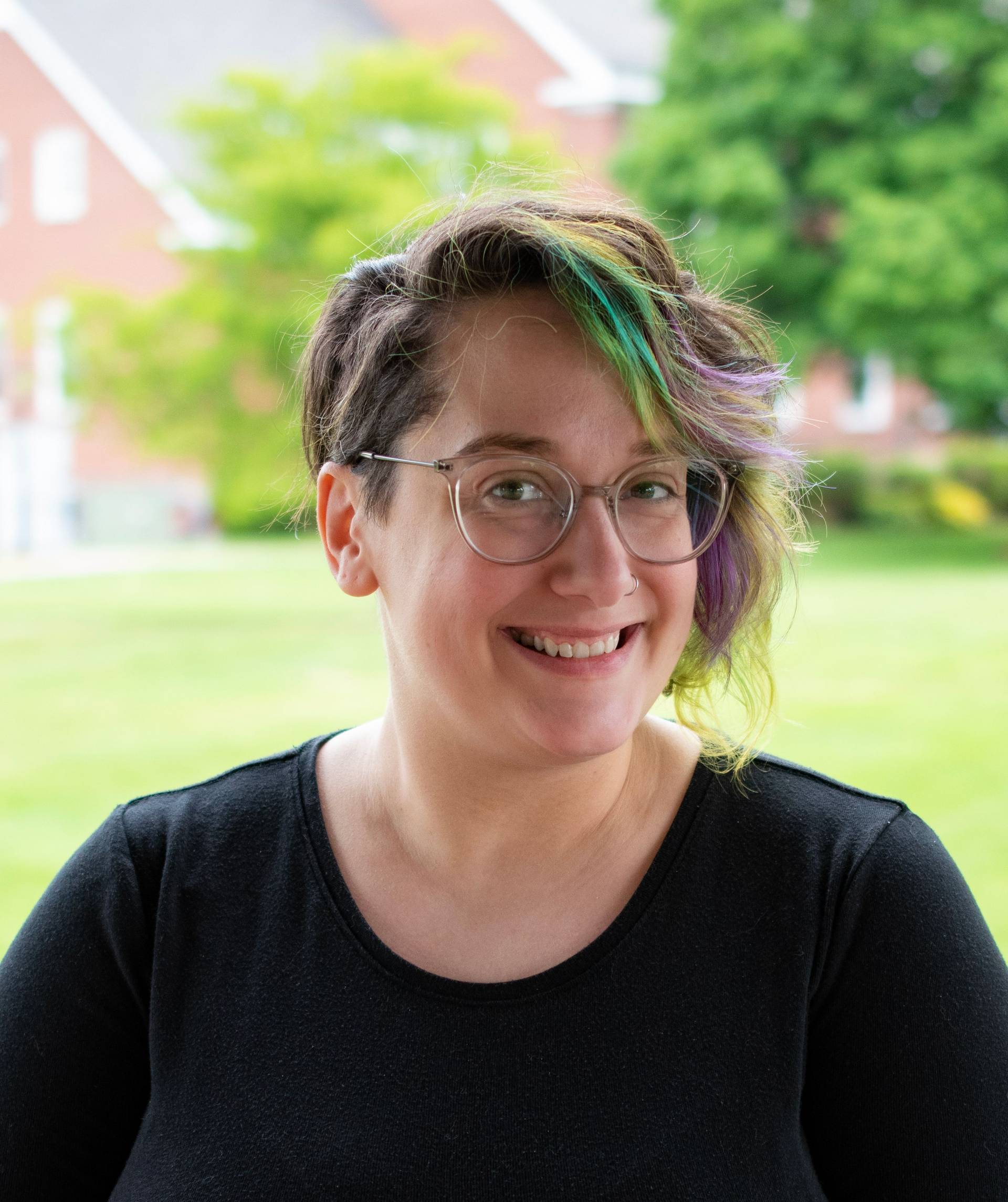 Woman with glasses, dyed hair smiles at the camera, outdoors.