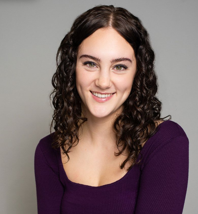 Young woman with dark curly hair smiles, wearing a purple top, against a gray backdrop.