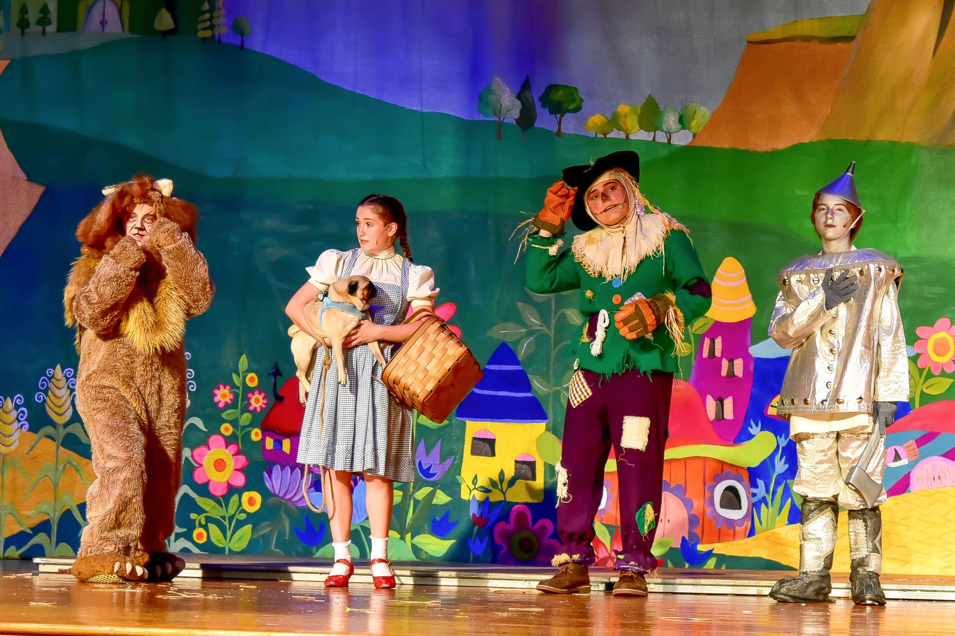A stage production of The Wizard of Oz: Lion, Dorothy, Scarecrow, Tin Man stand before painted backdrop.