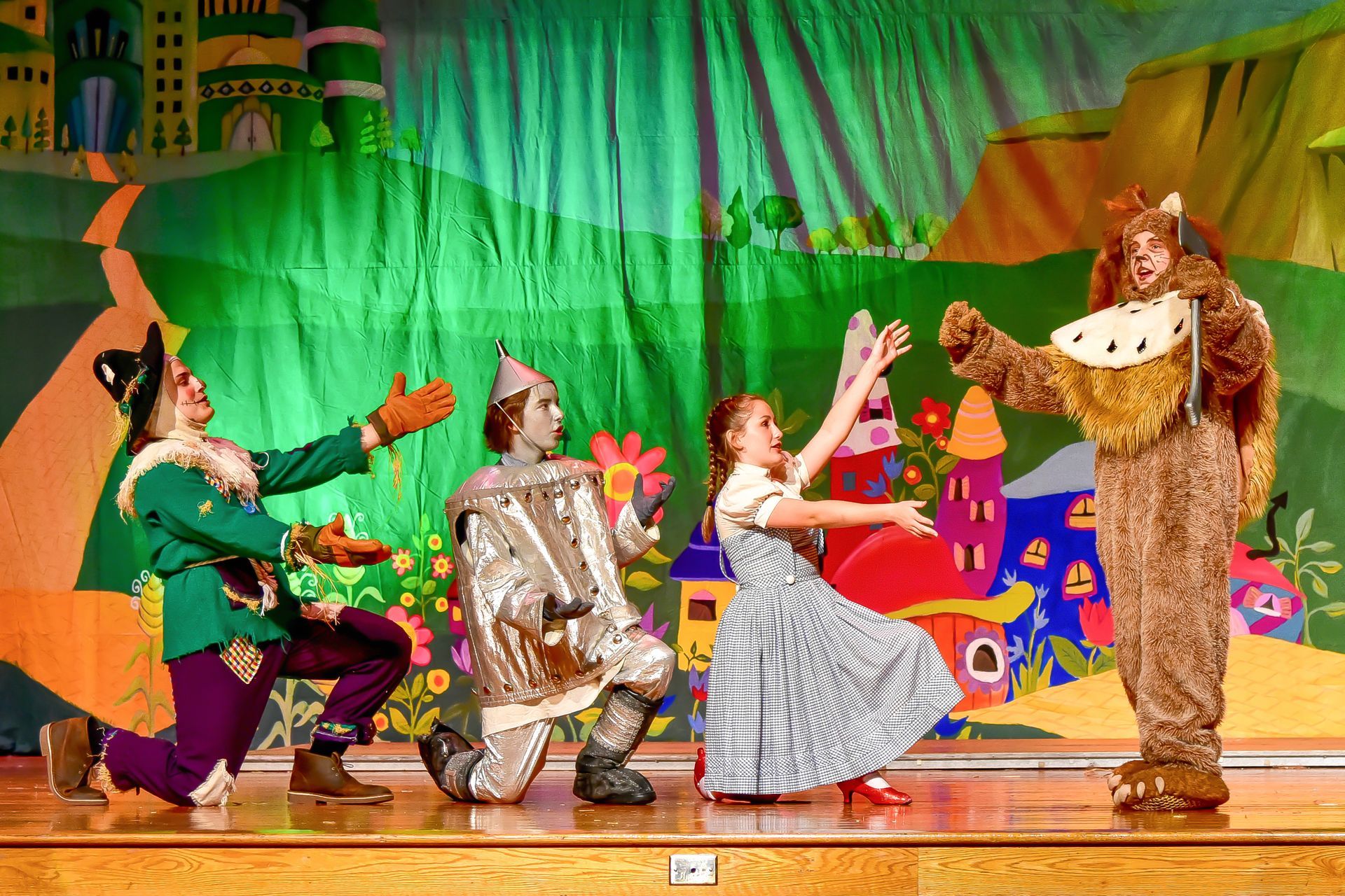 Actors in Wizard of Oz costumes on a stage: Scarecrow, Tin Man, Dorothy, and Cowardly Lion.