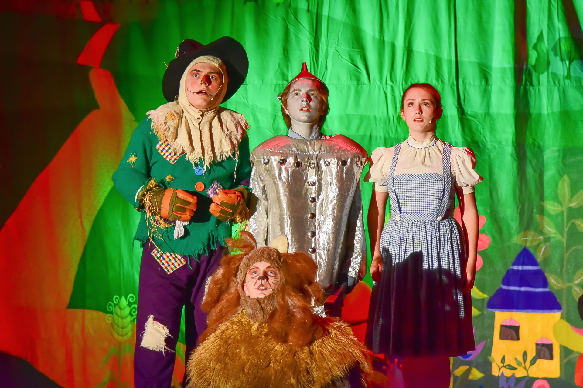 Group in costumes from The Wizard of Oz: Scarecrow, Tin Man, Dorothy, Cowardly Lion, green backdrop.