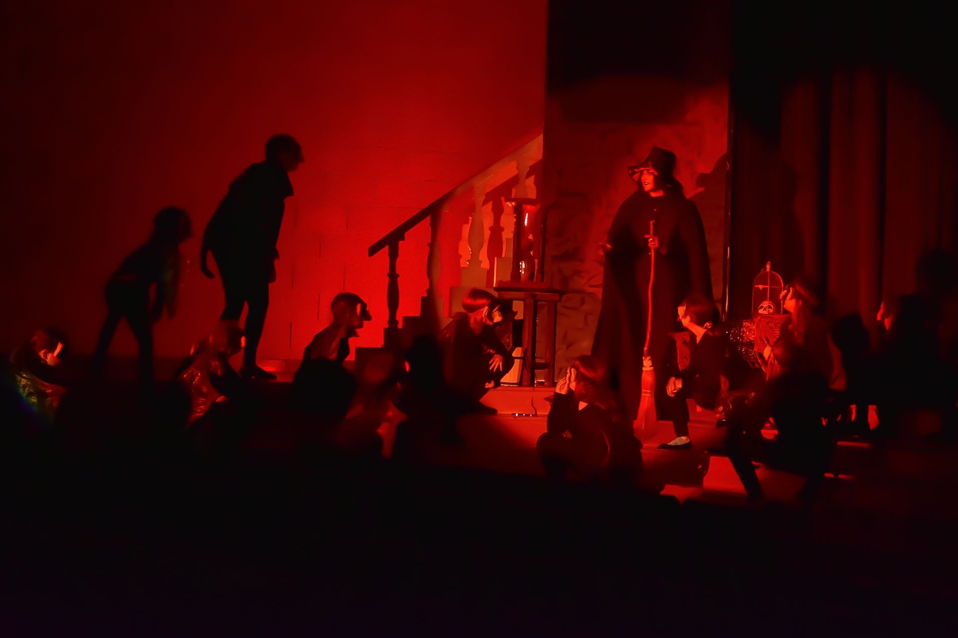 A stage with silhouetted figures under red light; one tall figure stands amidst a seated group.