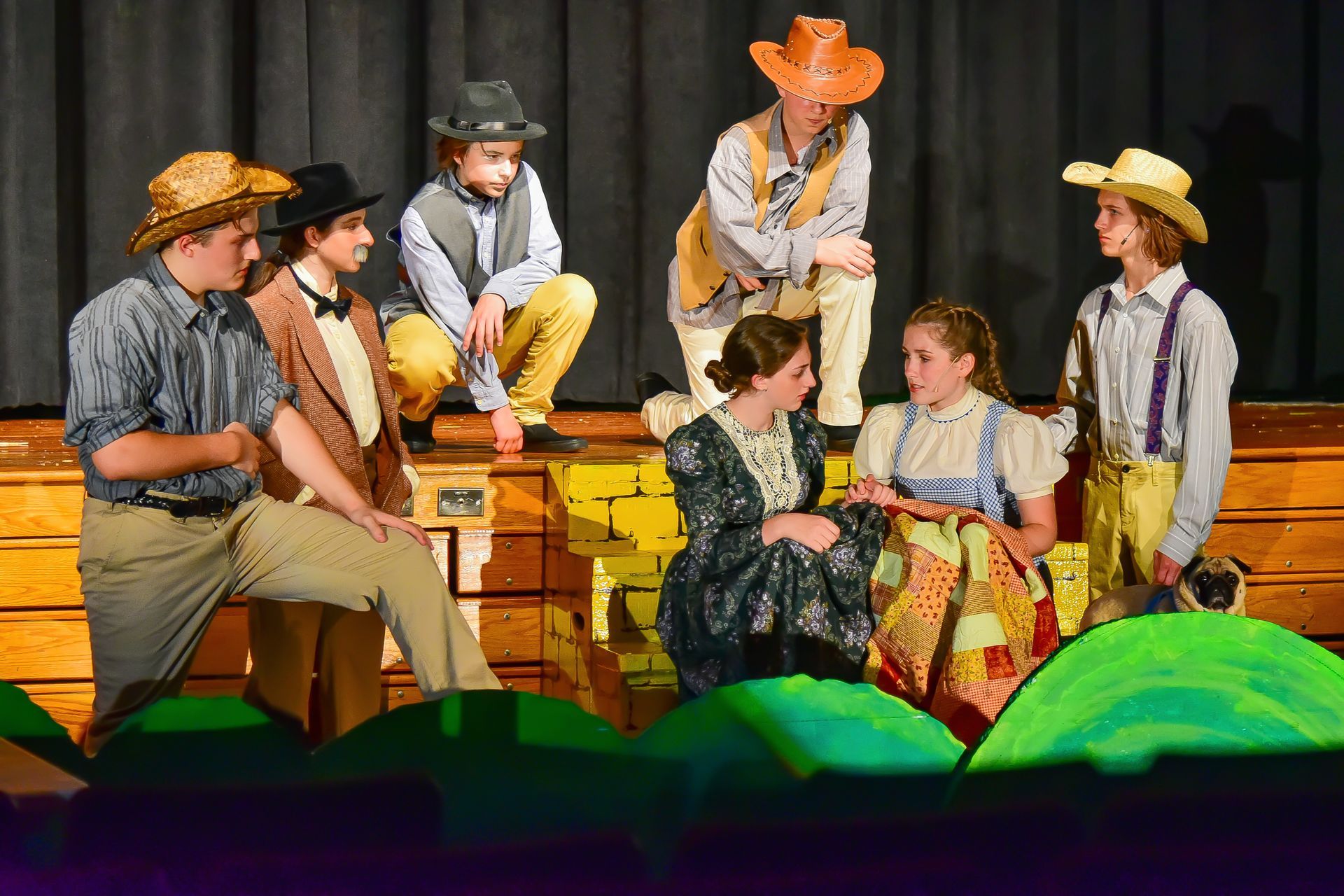 Actors in a play wearing costumes, on a stage with painted backdrop and set pieces.