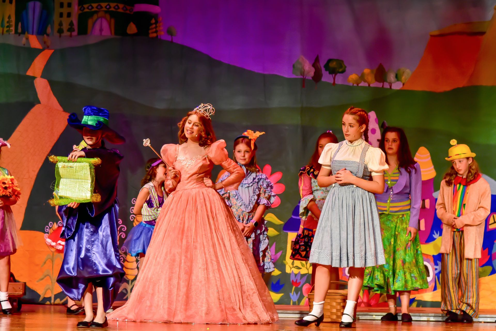 Stage production of The Wizard of Oz; characters on stage with colorful costumes, backdrop is a painted yellow brick road.