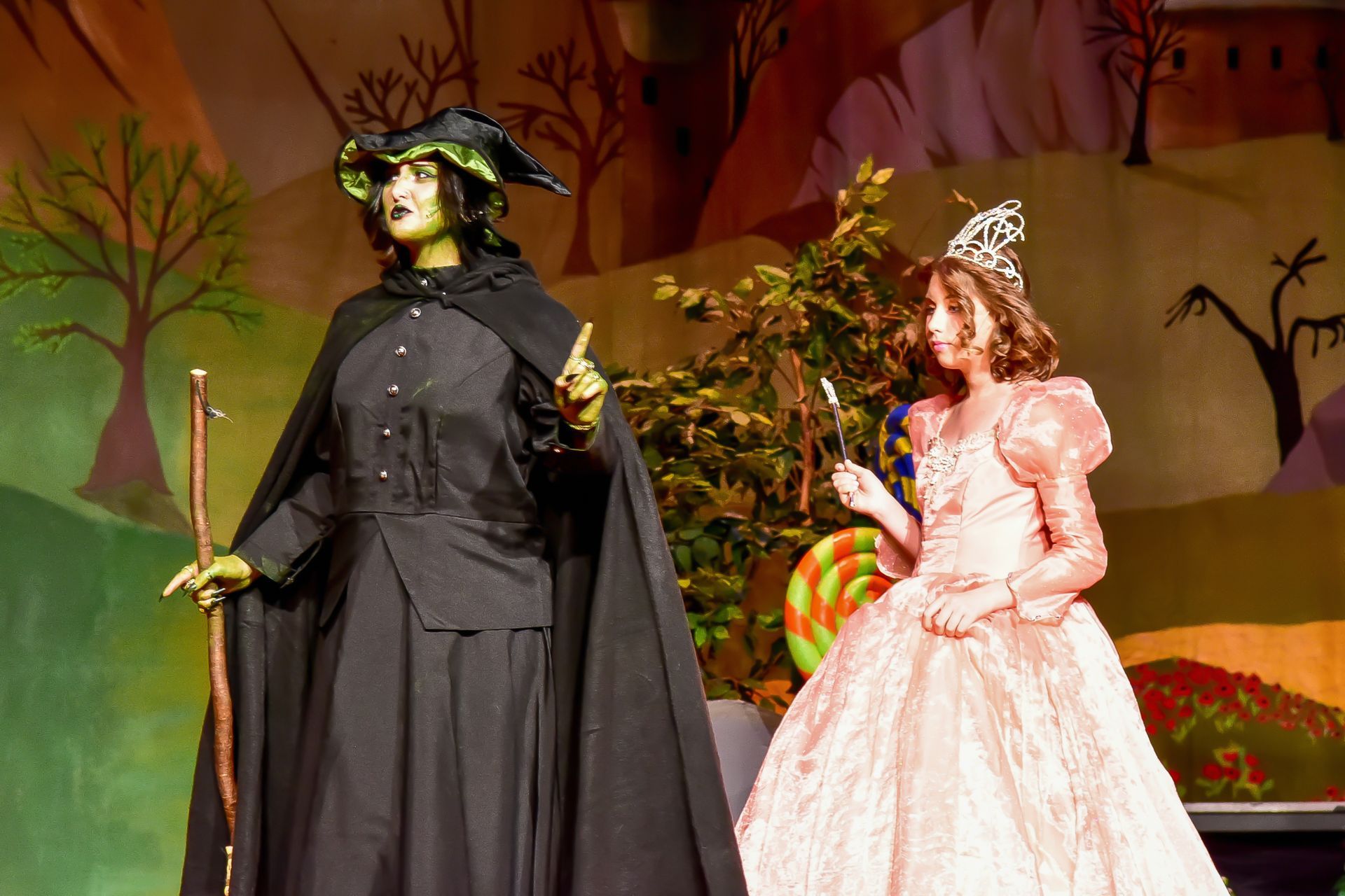 Wicked Witch of the West and a young girl in a pink dress on a stage set with a lollipop.