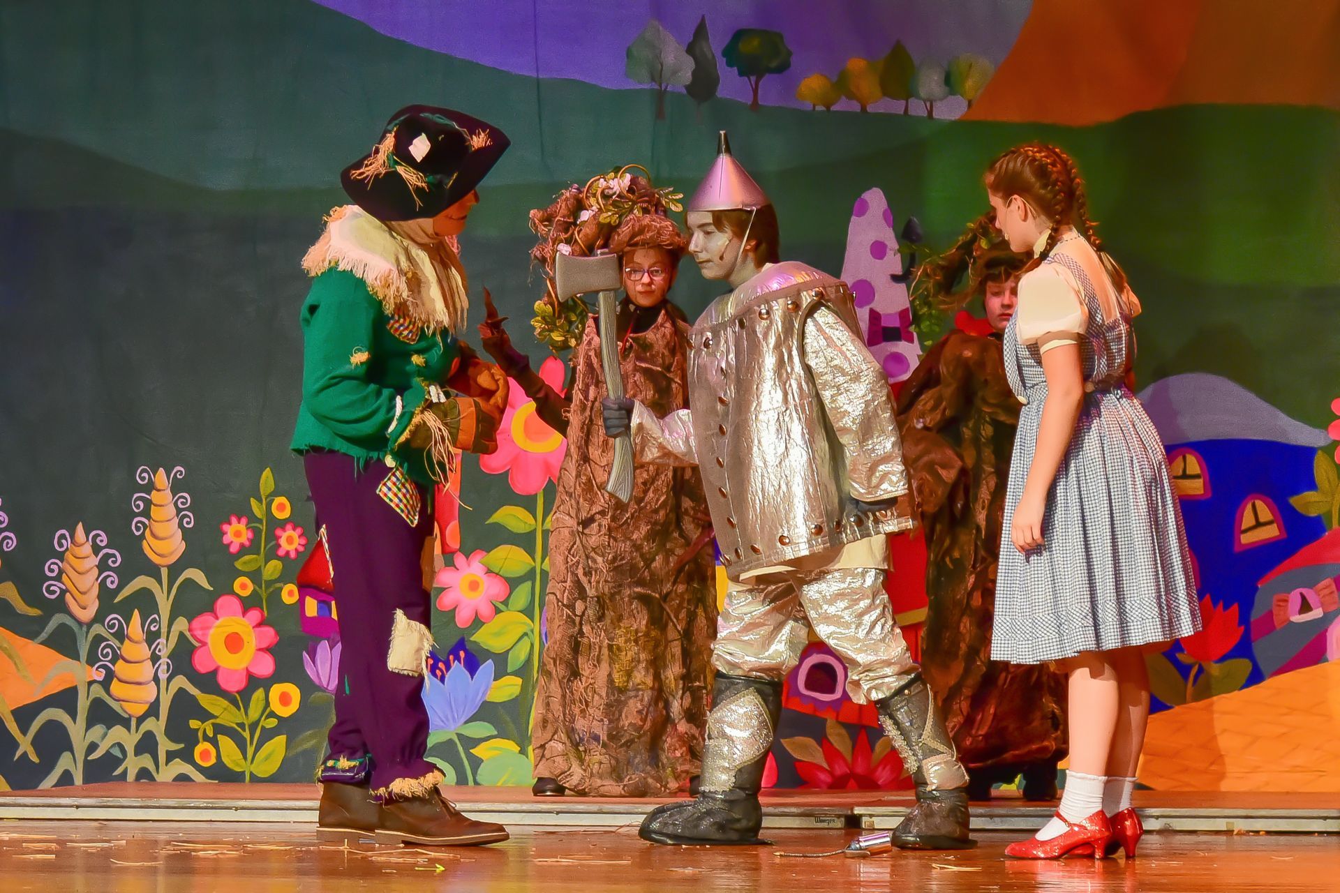 Actors in Wizard of Oz costumes on stage. Scarecrow, Tin Man, Dorothy, others. Colorful floral backdrop.