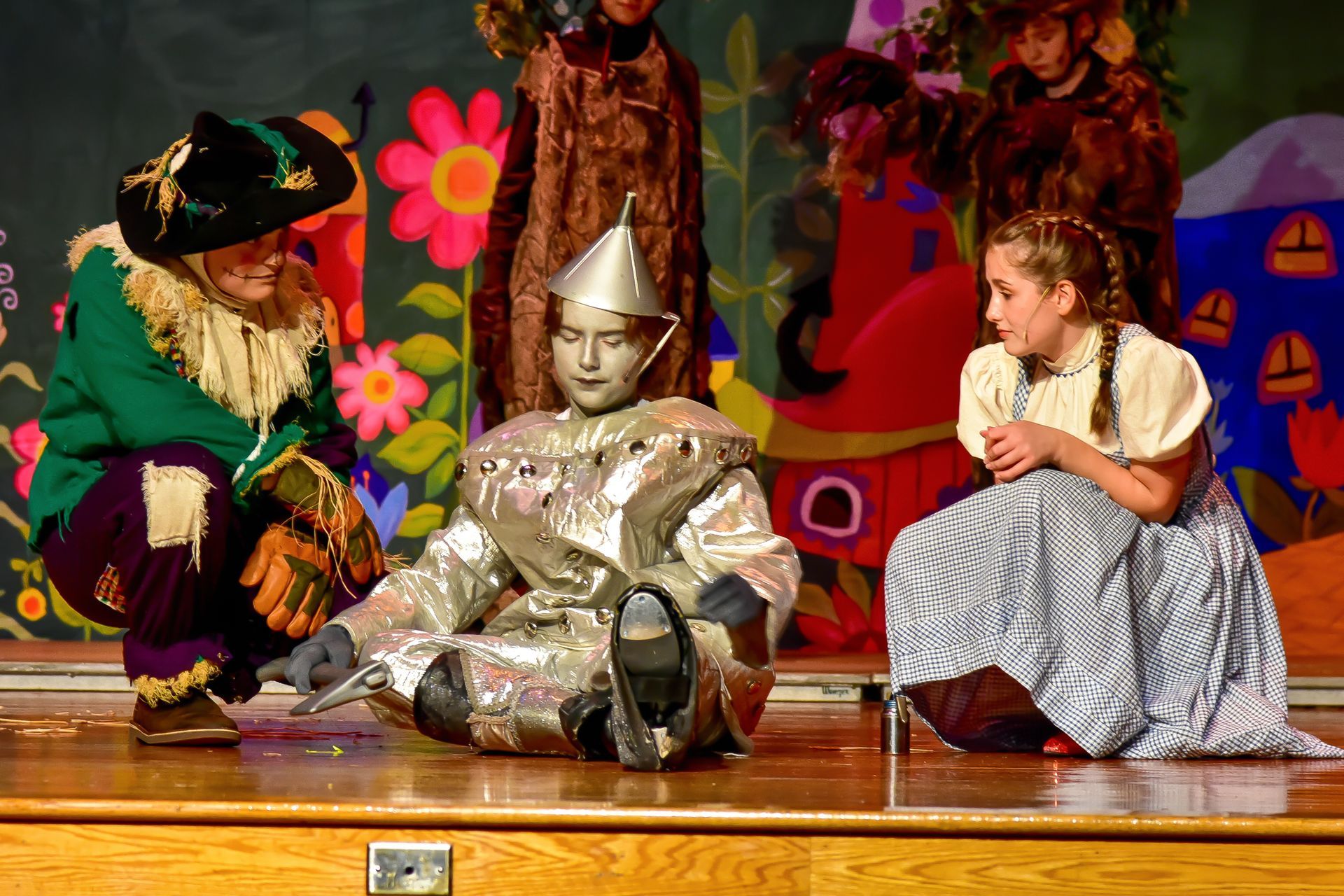 Actors in costumes on stage. Scarecrow, Tin Man, and Dorothy seated; background of Oz scene.