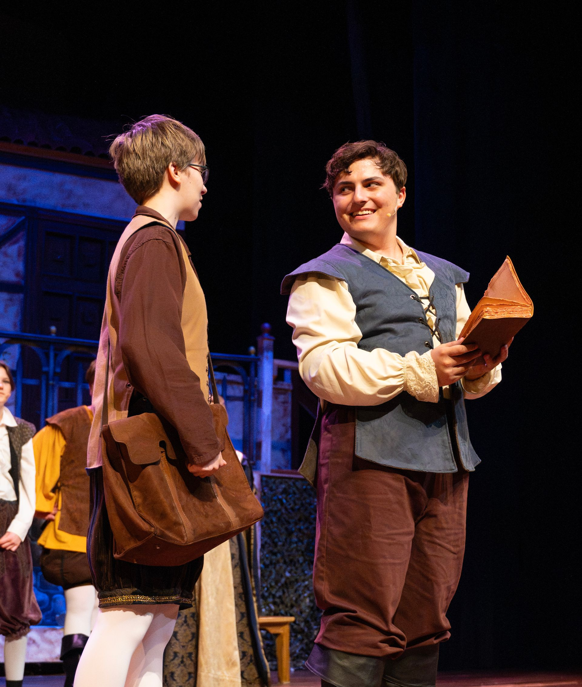 Two actors in period costumes on stage, one holding a book, smiling, the other facing them.