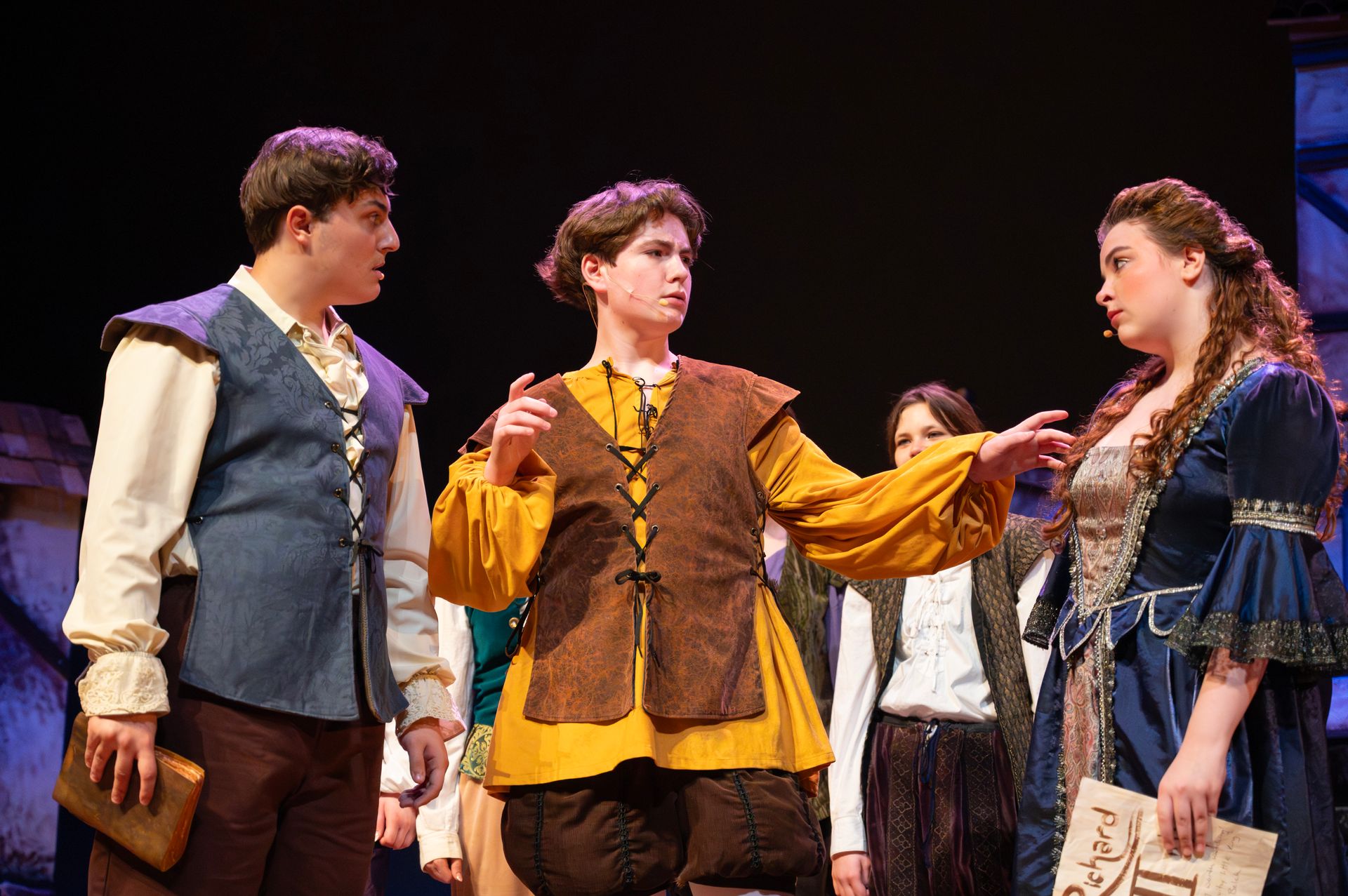 Three costumed actors on stage, performing. Man in yellow gestures, two others listen, looking concerned.