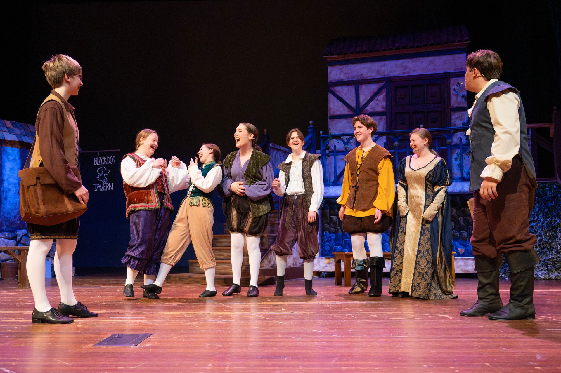A stage production: actors in Renaissance costumes on a wooden stage, laughing and interacting.
