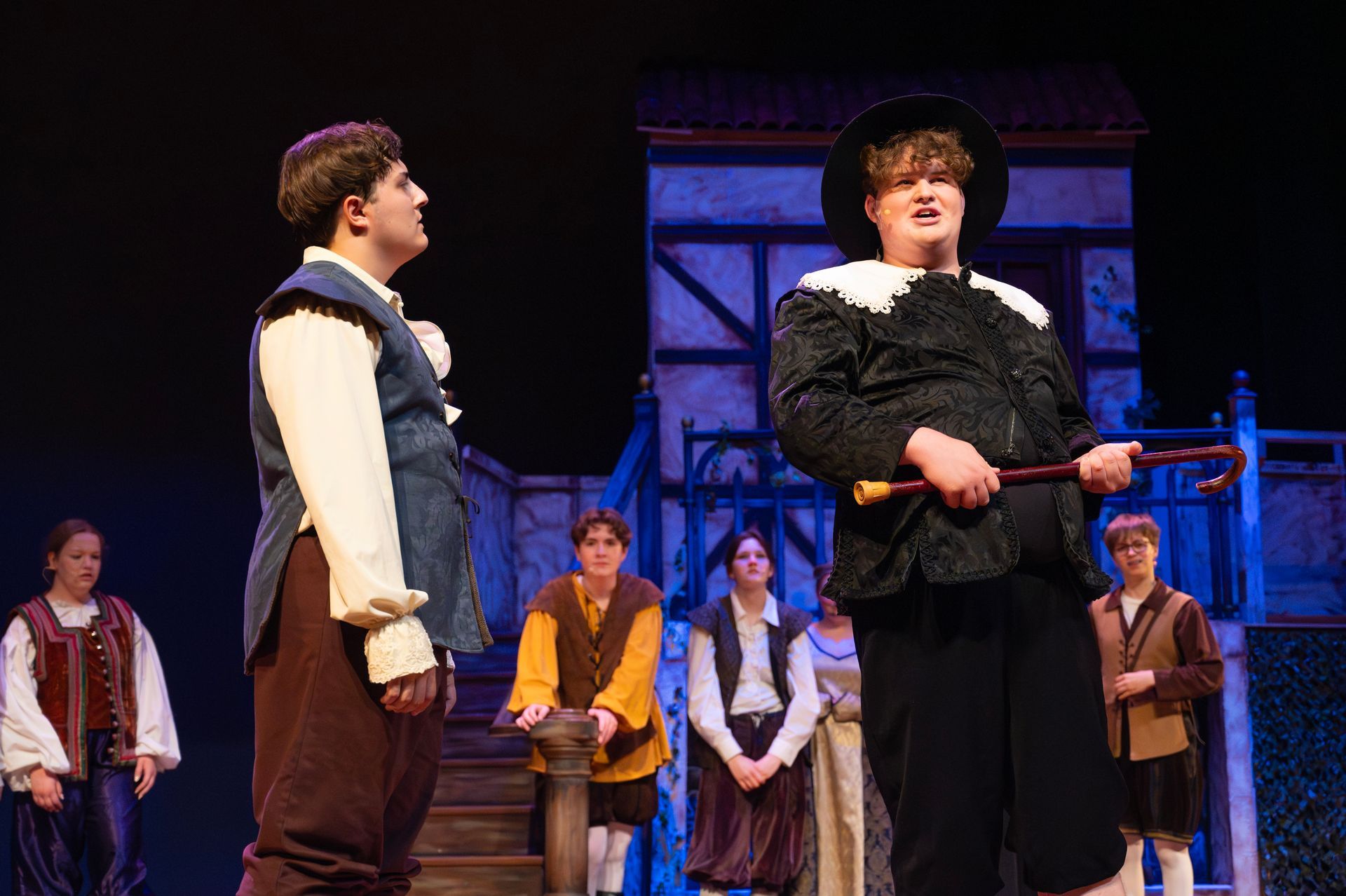 Actors on a stage, some in period costumes, performing in front of a backdrop.
