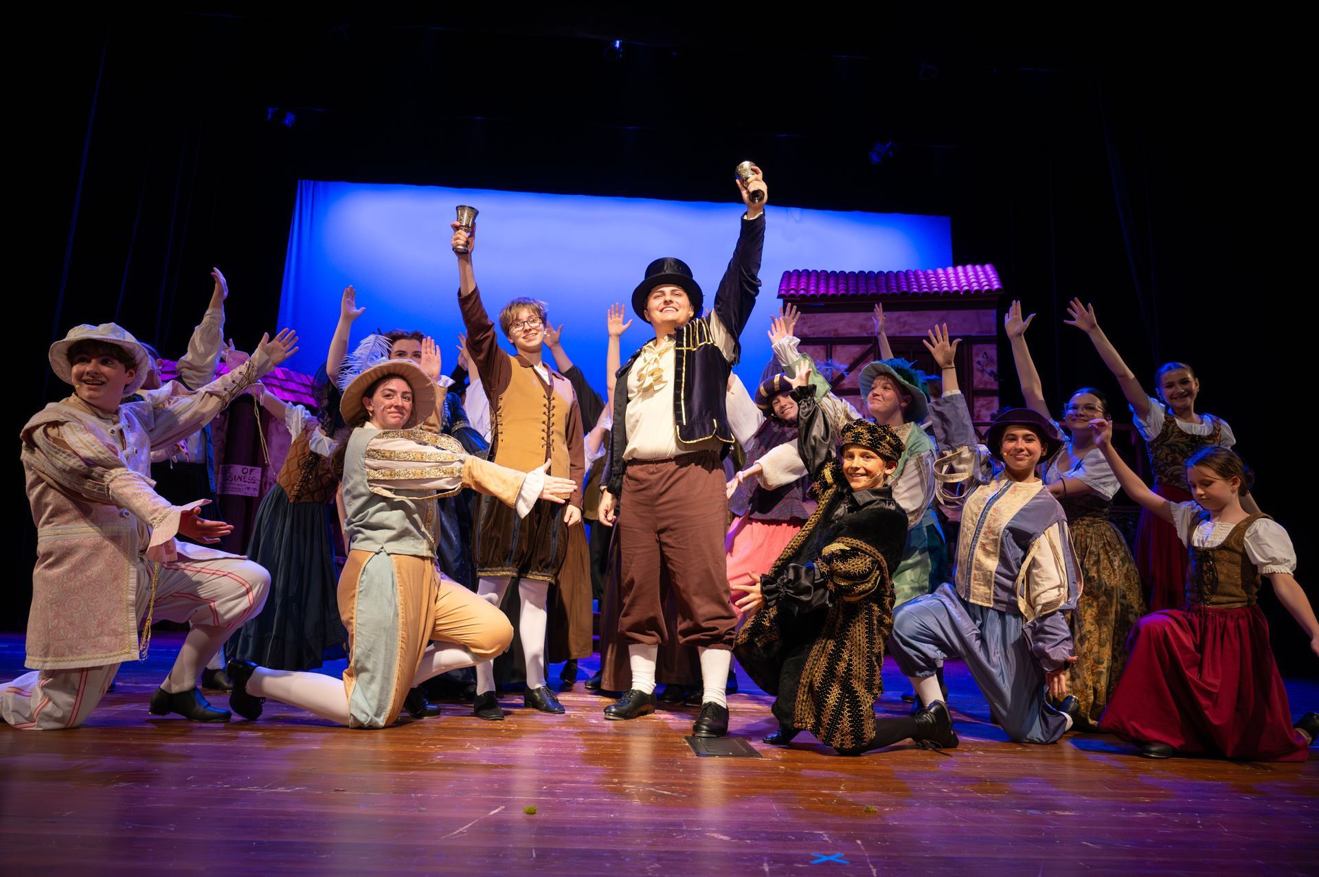 A costumed cast on stage raises arms. Bright stage lighting.