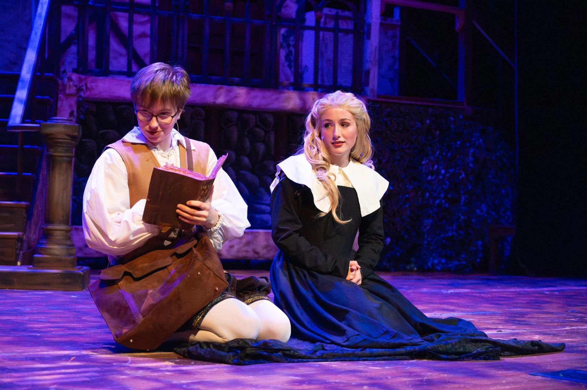 Two people in period costumes on a stage. A man reads a book, while a woman sits.