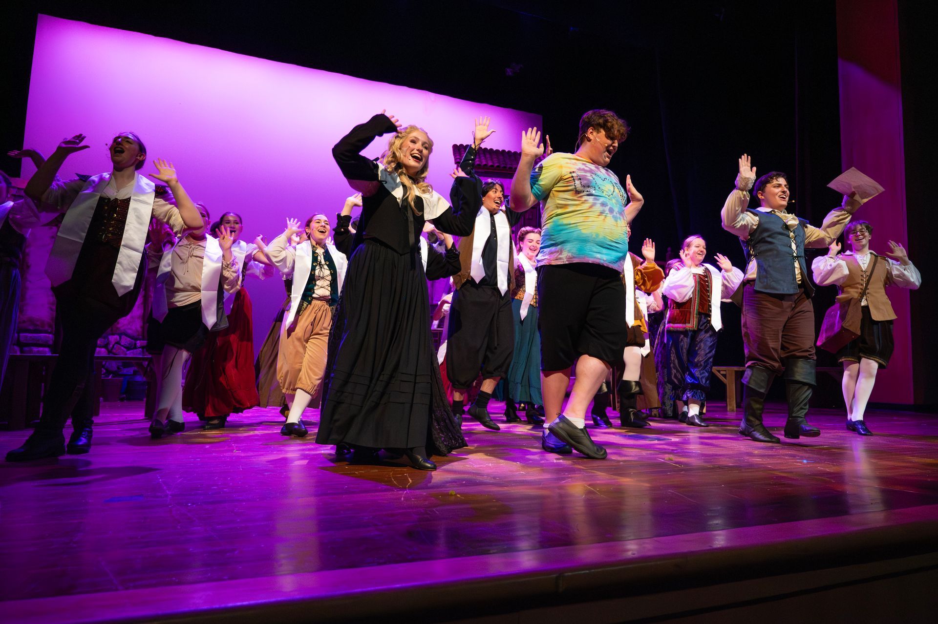 A stage production with actors in costume. A person in tie-dye stands out. Purple stage lights.