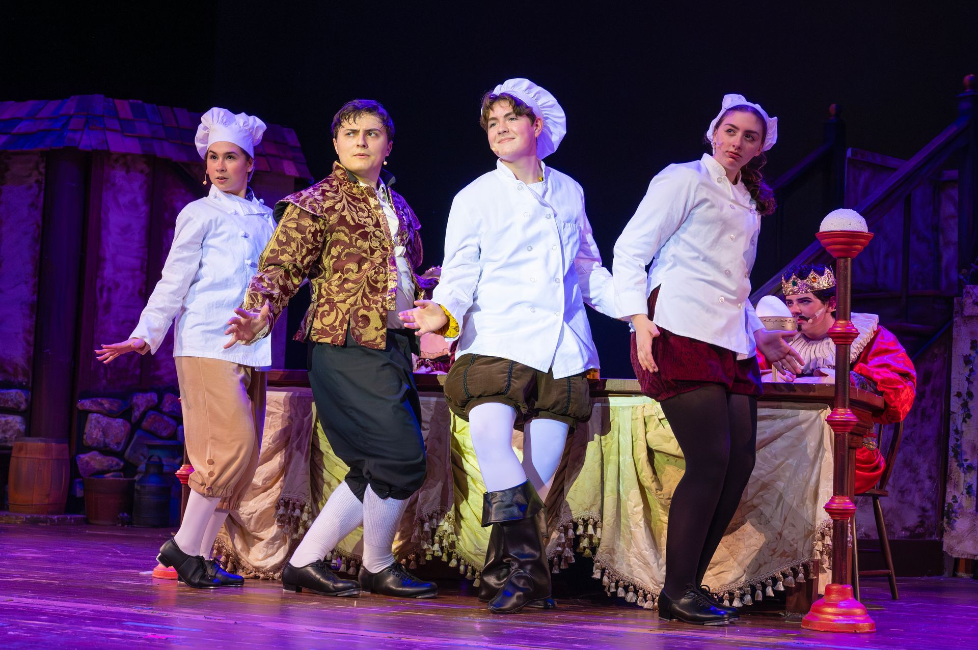 Four actors in chef hats dance onstage, next to a seated king. Stage is rustic and purple lit.