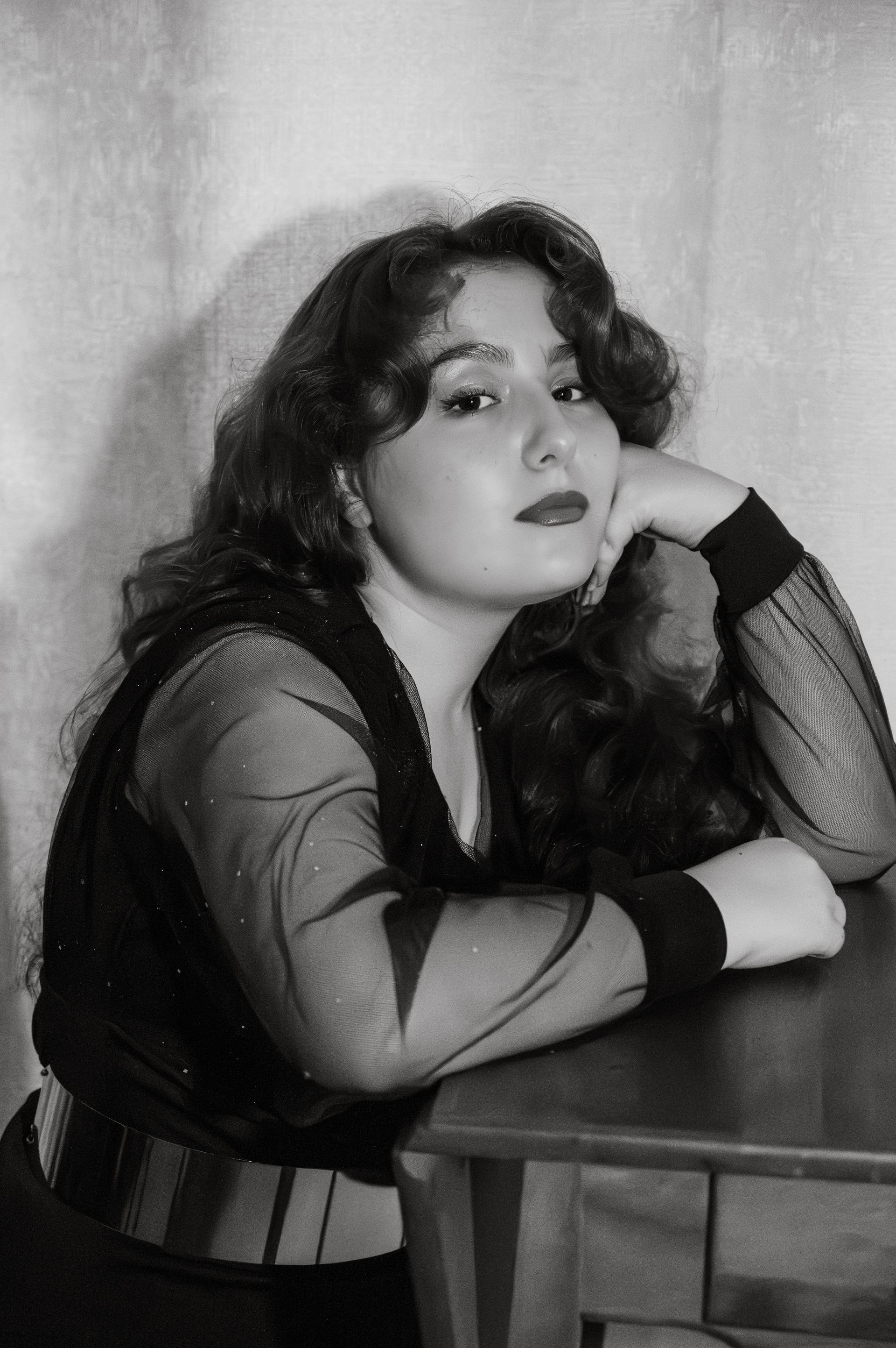 Woman in black outfit, leaning on table. Black and white photo, serious expression.