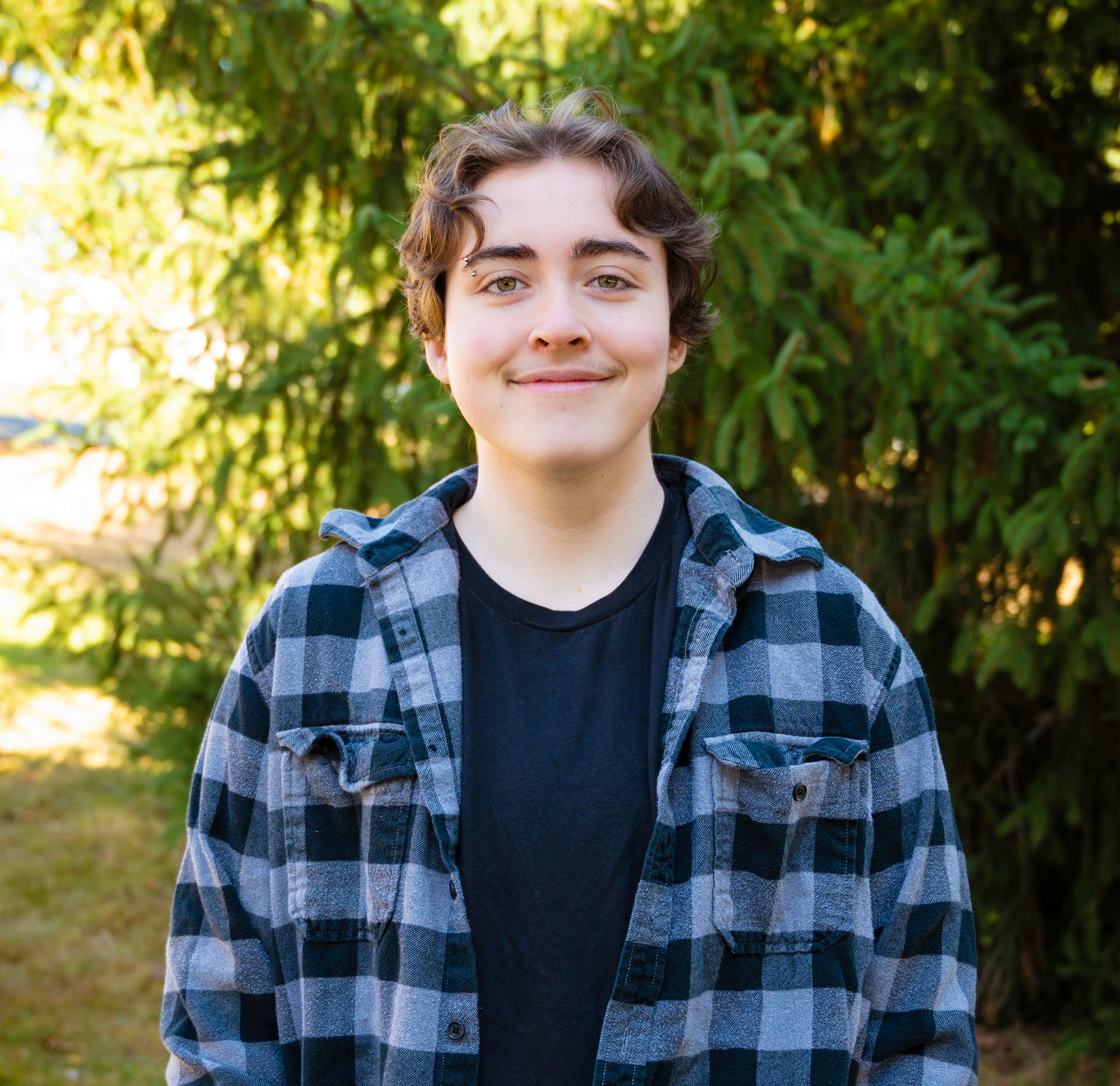 Young person with curly brown hair, wearing a black shirt and plaid jacket, smiling outdoors.