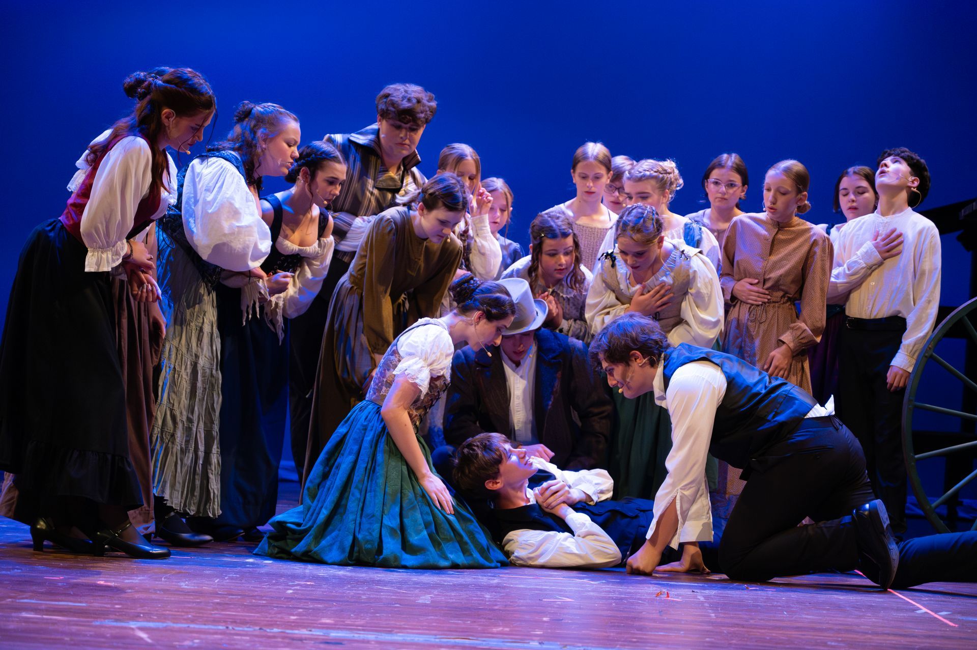 A group of people in period clothing surround a person on stage. Dark blue background.