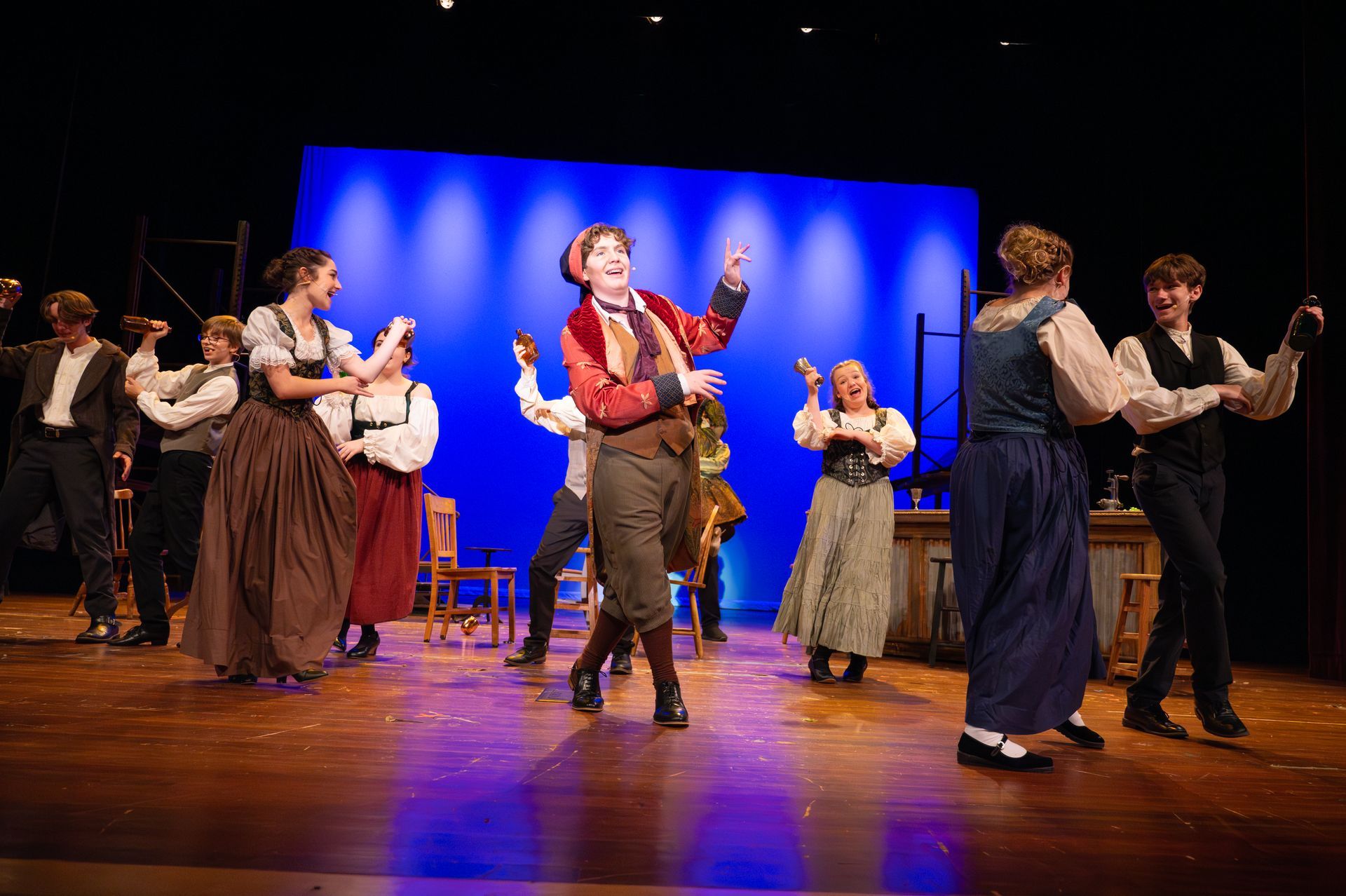 A stage performance with dancers in traditional costumes, with a blue backdrop, wood stage, and chairs.