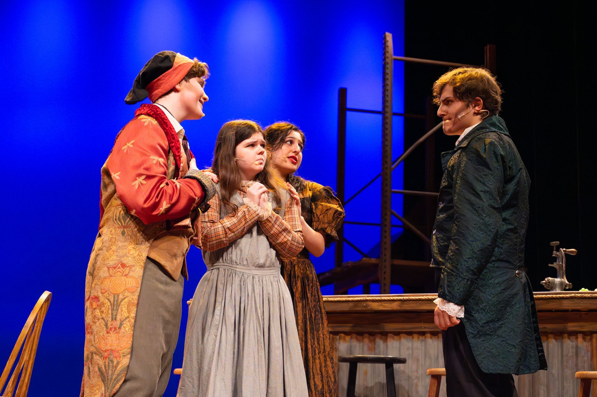 Four costumed actors on a stage: two men, two women. They appear to be in a play. Blue backdrop.