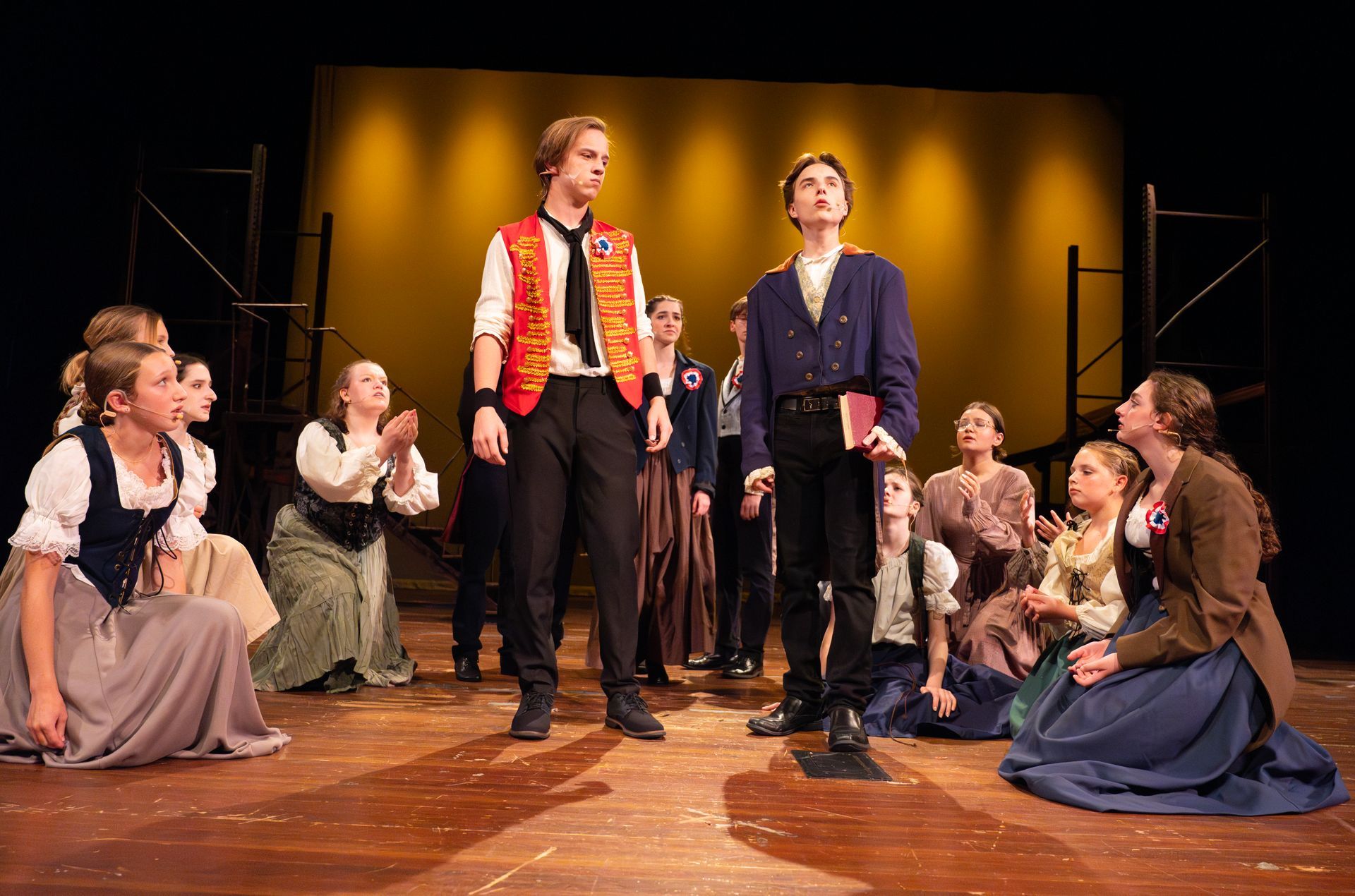 Actors in period costumes on stage; two young men stand, others kneel, watch.