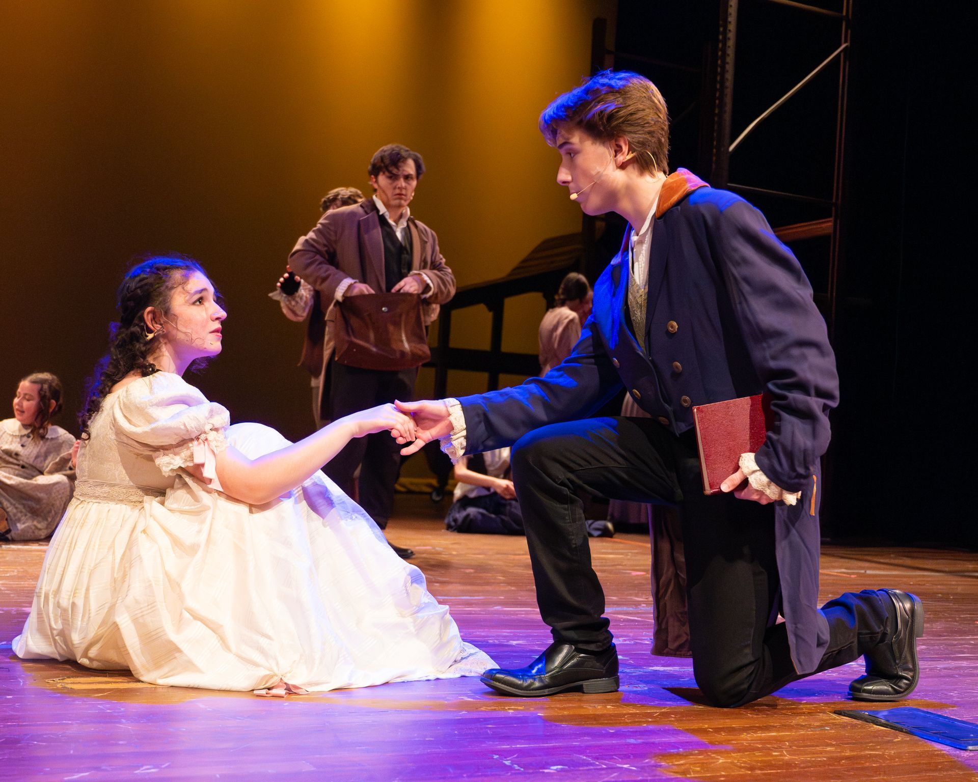 Actors in period costumes perform on stage: Man kneels to woman, holding her hand. Other actors in background.