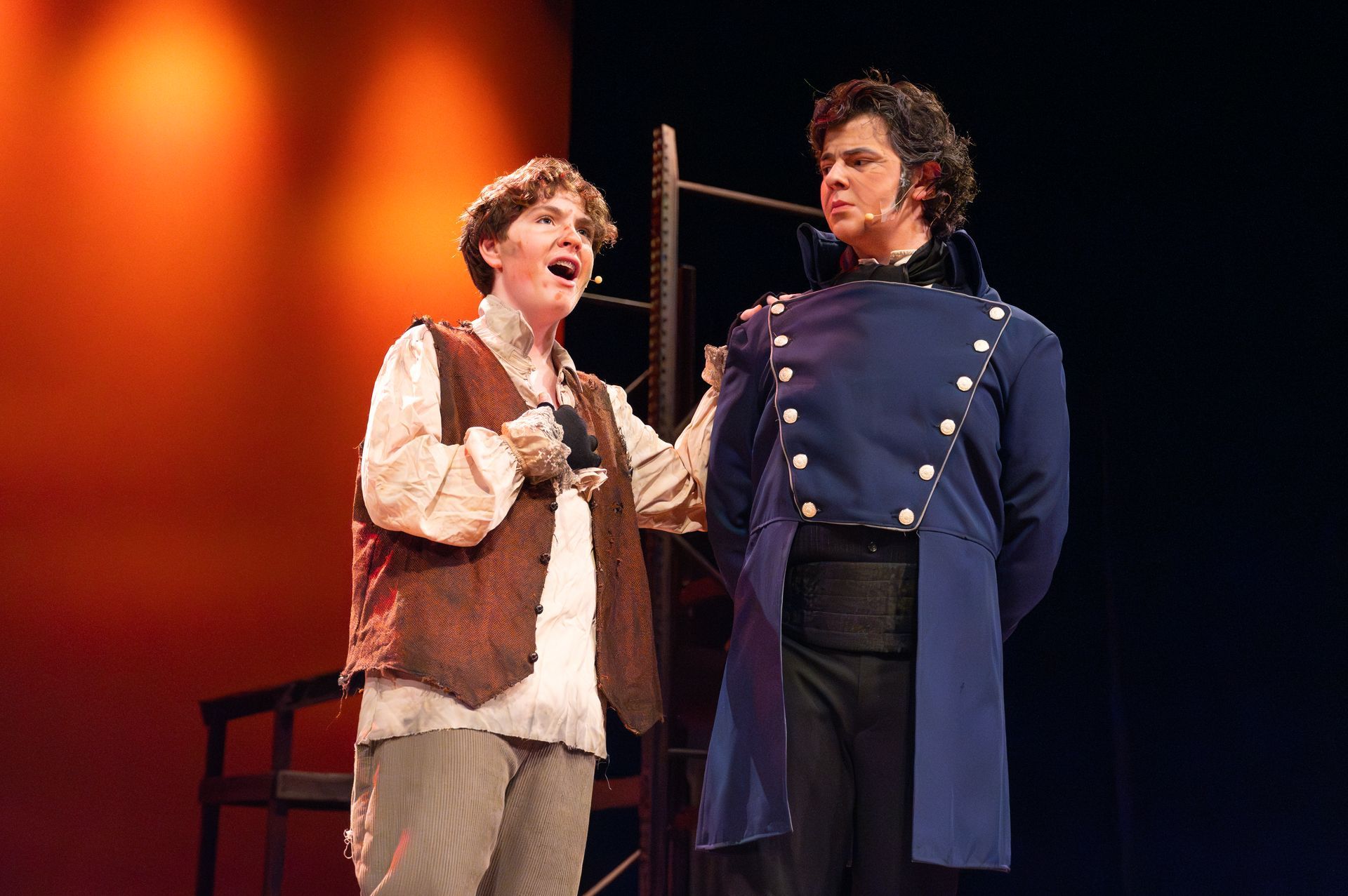Two men on stage, one in a vest singing, the other in a blue coat, listening. Orange backdrop.