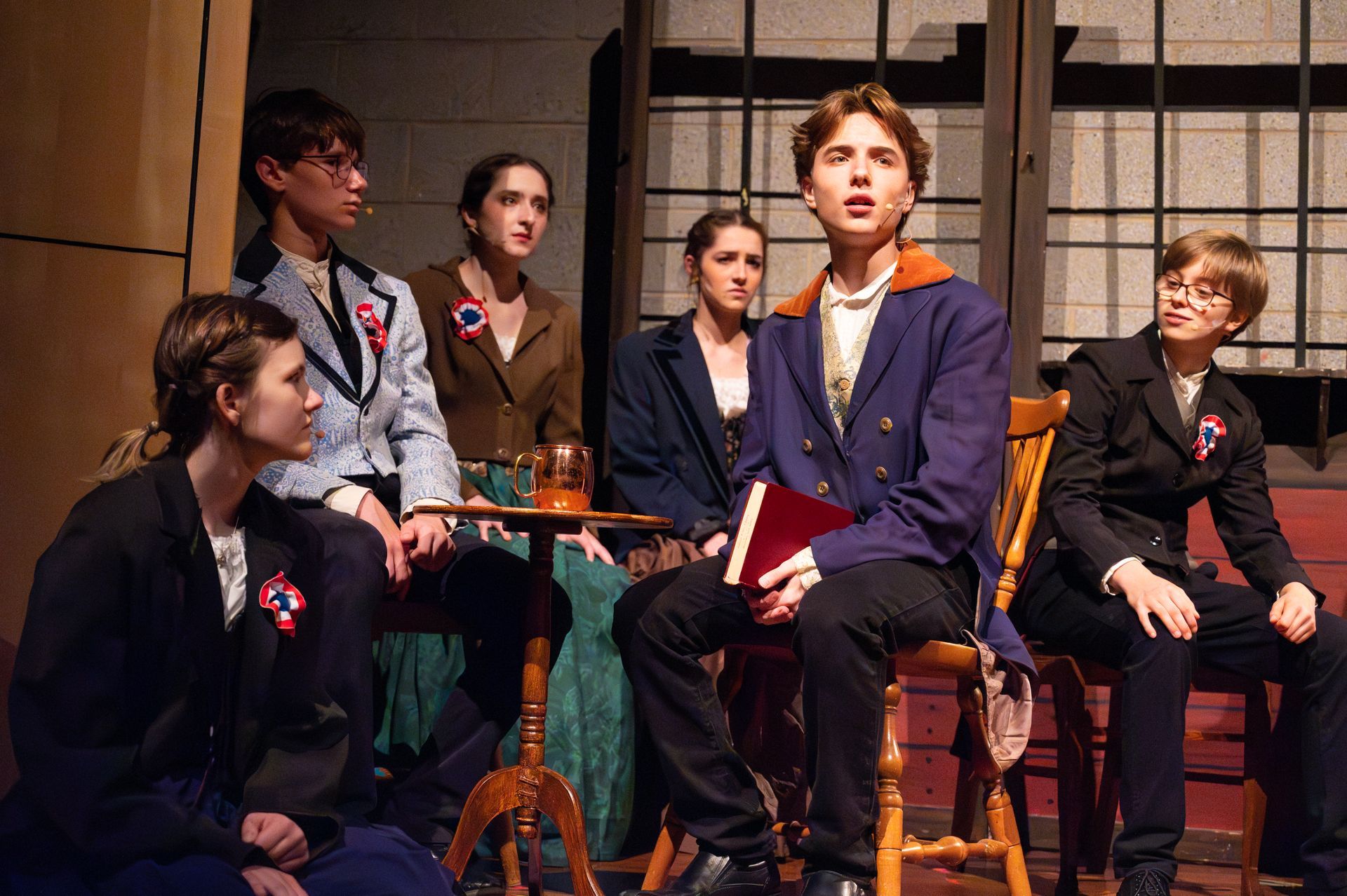 A play scene: Group of young actors in period costumes, some seated, one holding a book, all looking up.