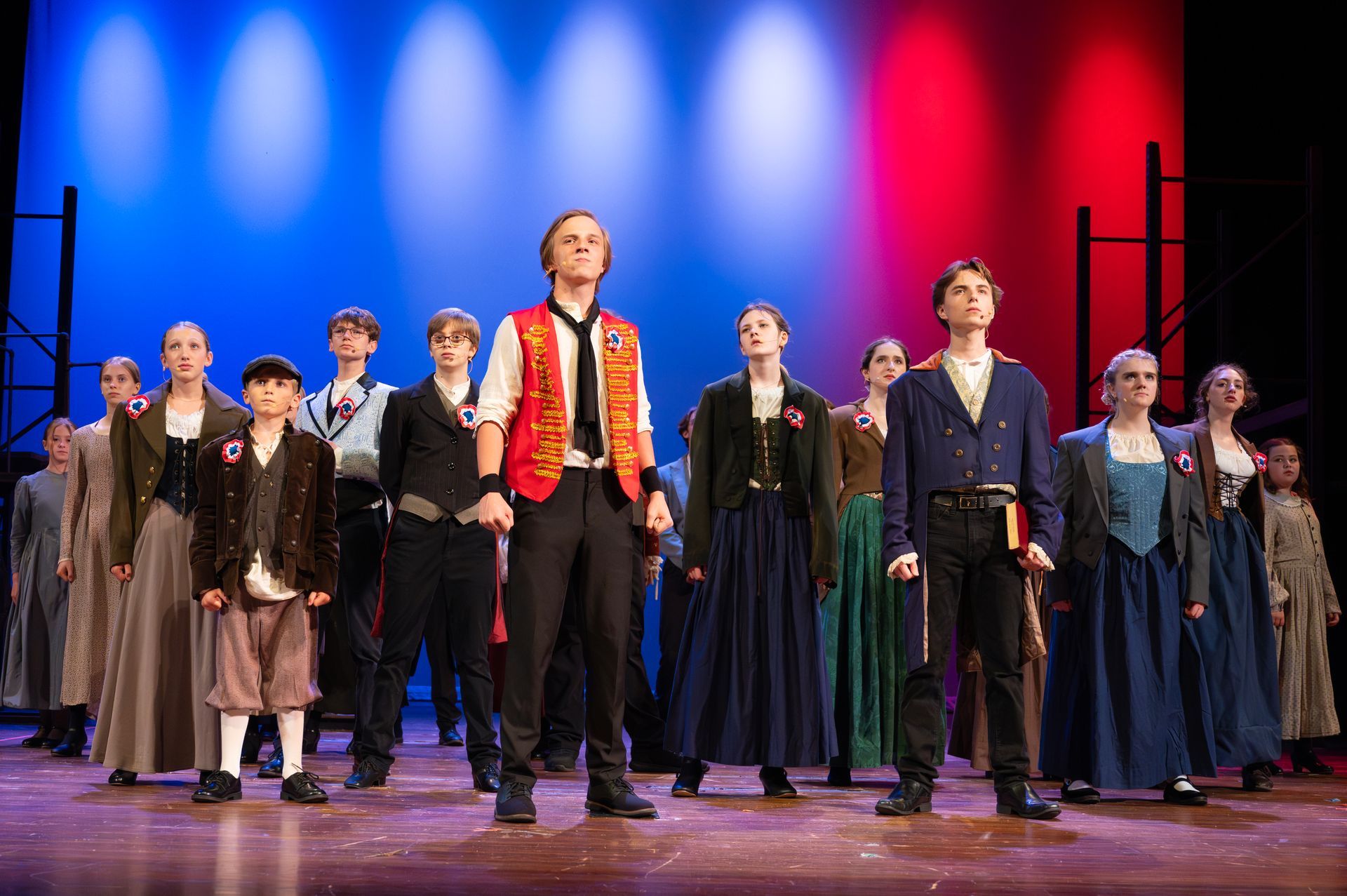 A group of people on stage in costumes, singing with red and blue stage lighting.