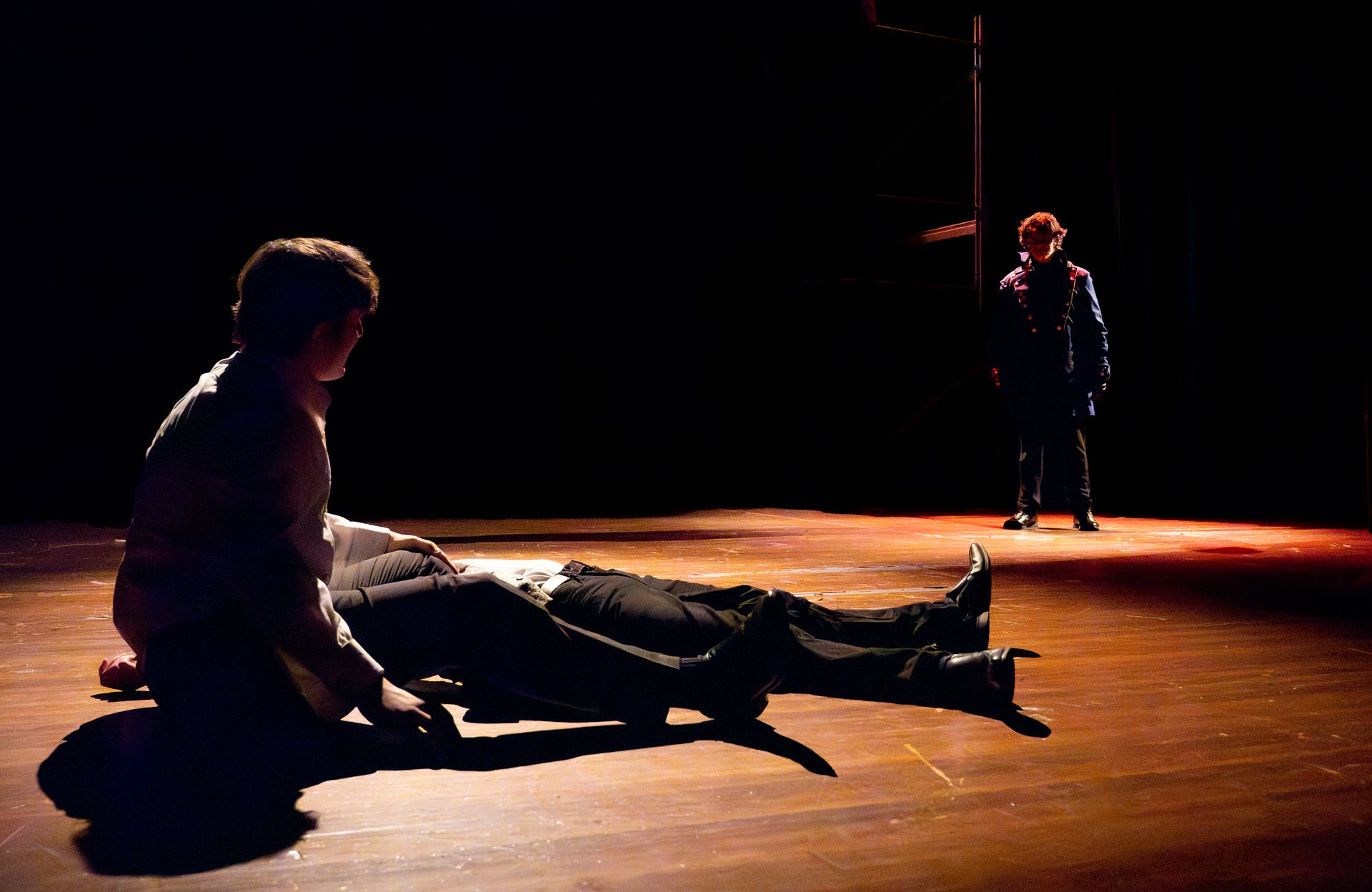On a dark stage, a person kneels by another lying on the floor. A third person stands in the background.