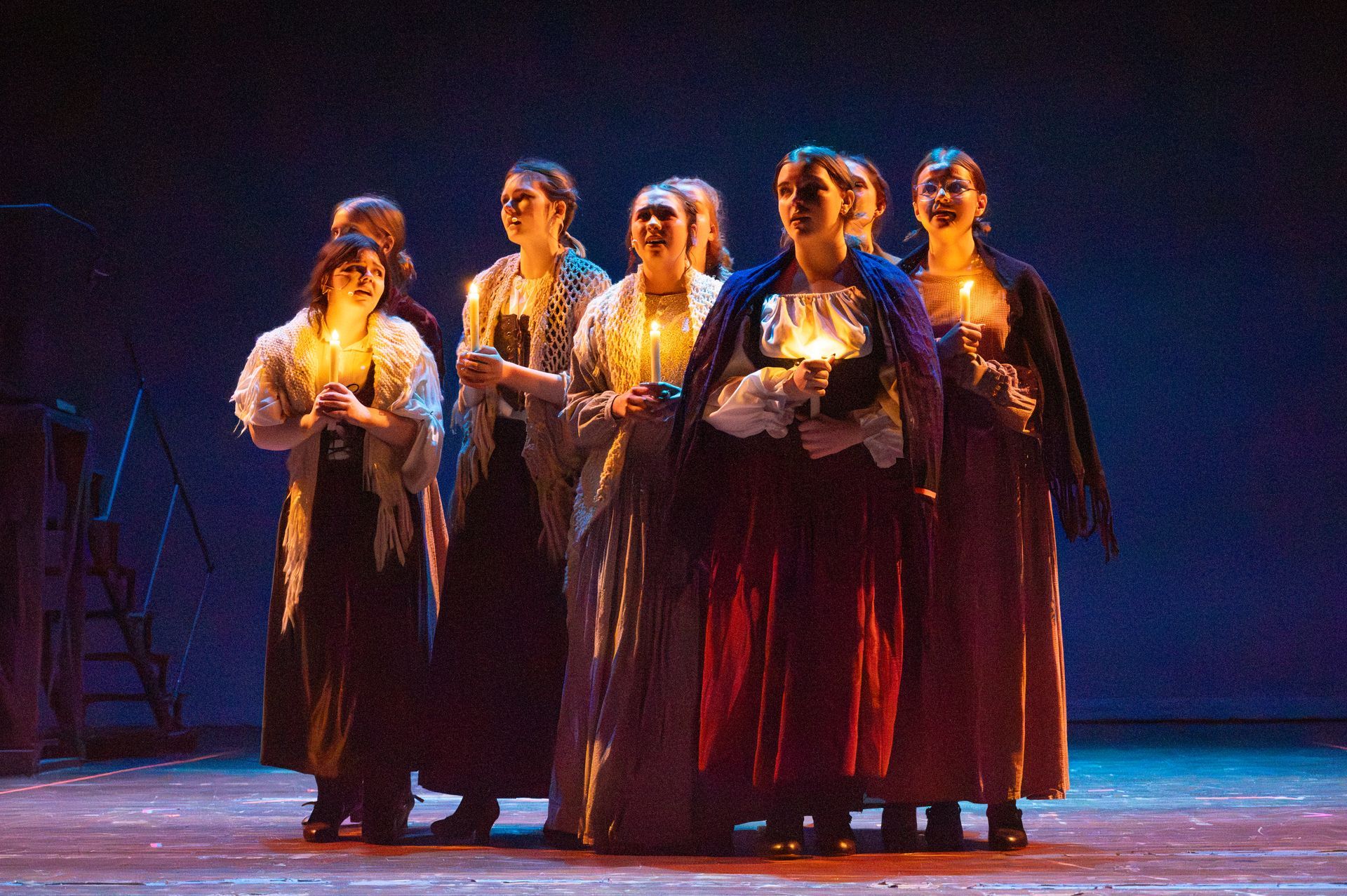 Group of women on stage in period costumes, holding candles, illuminated, dramatic lighting.