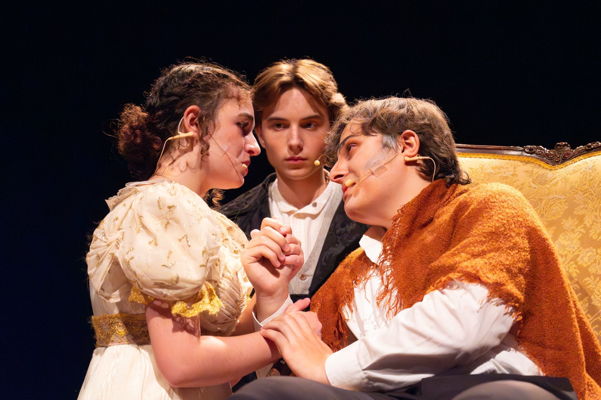 Three actors on stage: a woman in a white dress, a man in a brown poncho, and another in a black coat.
