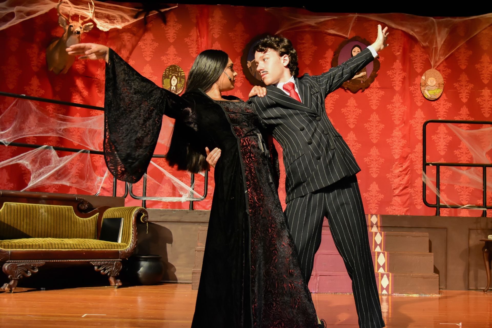 Actors in costume dancing on a stage. Woman in black gown, man in pinstripes. Red backdrop with cobwebs.