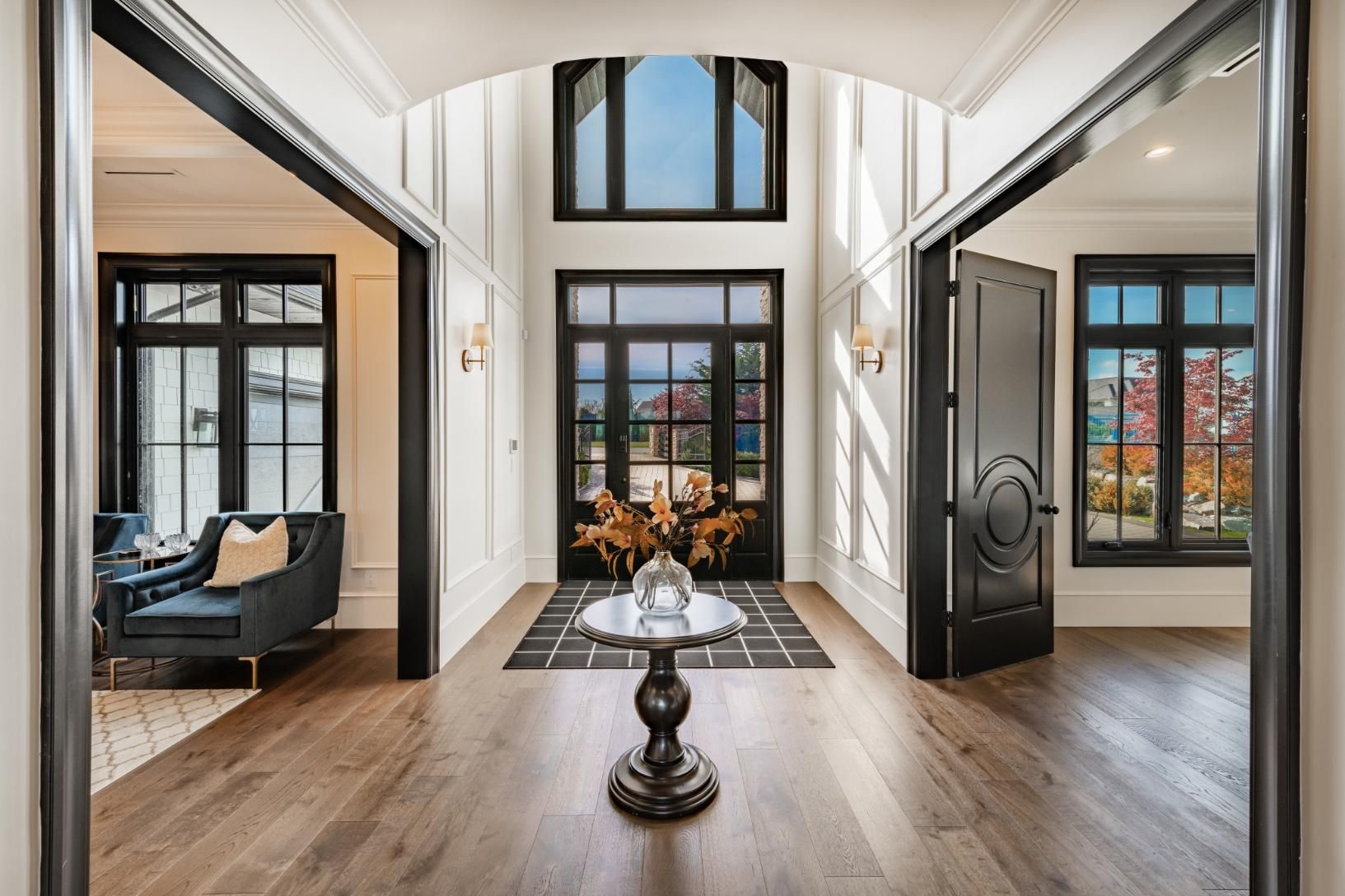 Elegant foyer with hardwood floor, black-framed windows/doors, and a central table.