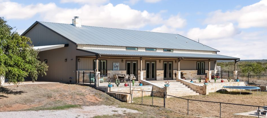 A long, light brown ranch-style house with a metal roof and a porch with a stone patio, set on a grassy field.