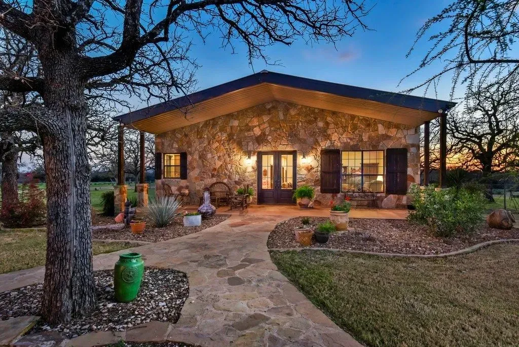 Stone house with covered porch, lit windows, and a walkway, at dusk.