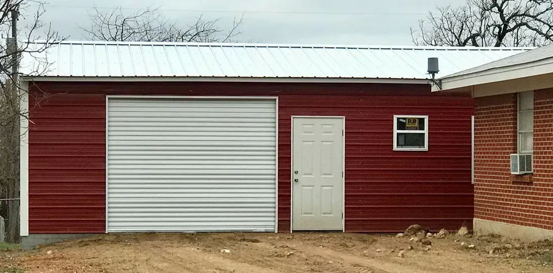 Red metal building with a white garage door, entry door, and window. White metal roof. Brick building on the right.