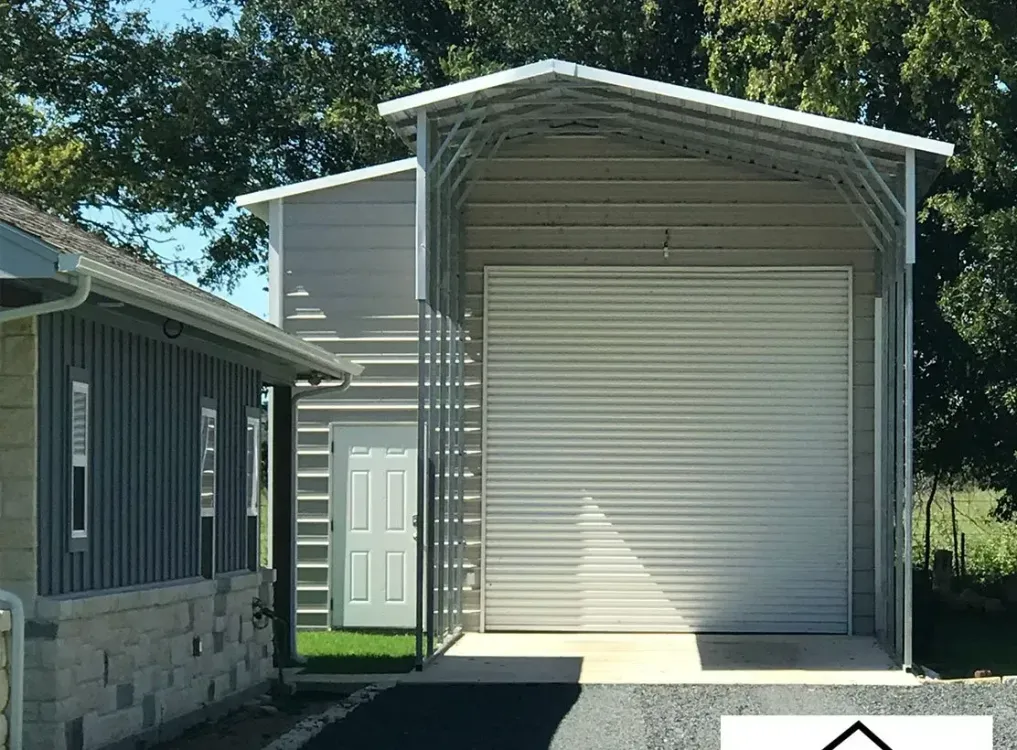 Metal carport with a large roll-up door, attached to a light-colored building with a white door.