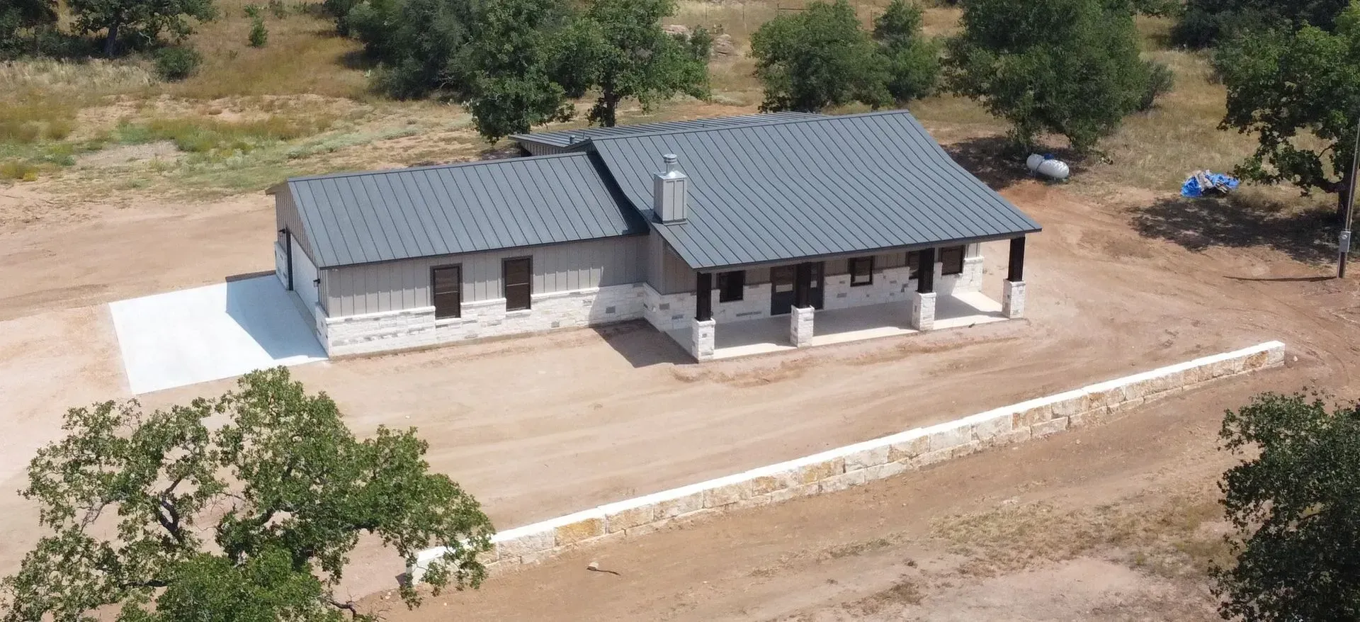 Aerial view of a stone house with a gray metal roof, surrounded by trees and a gravel driveway.