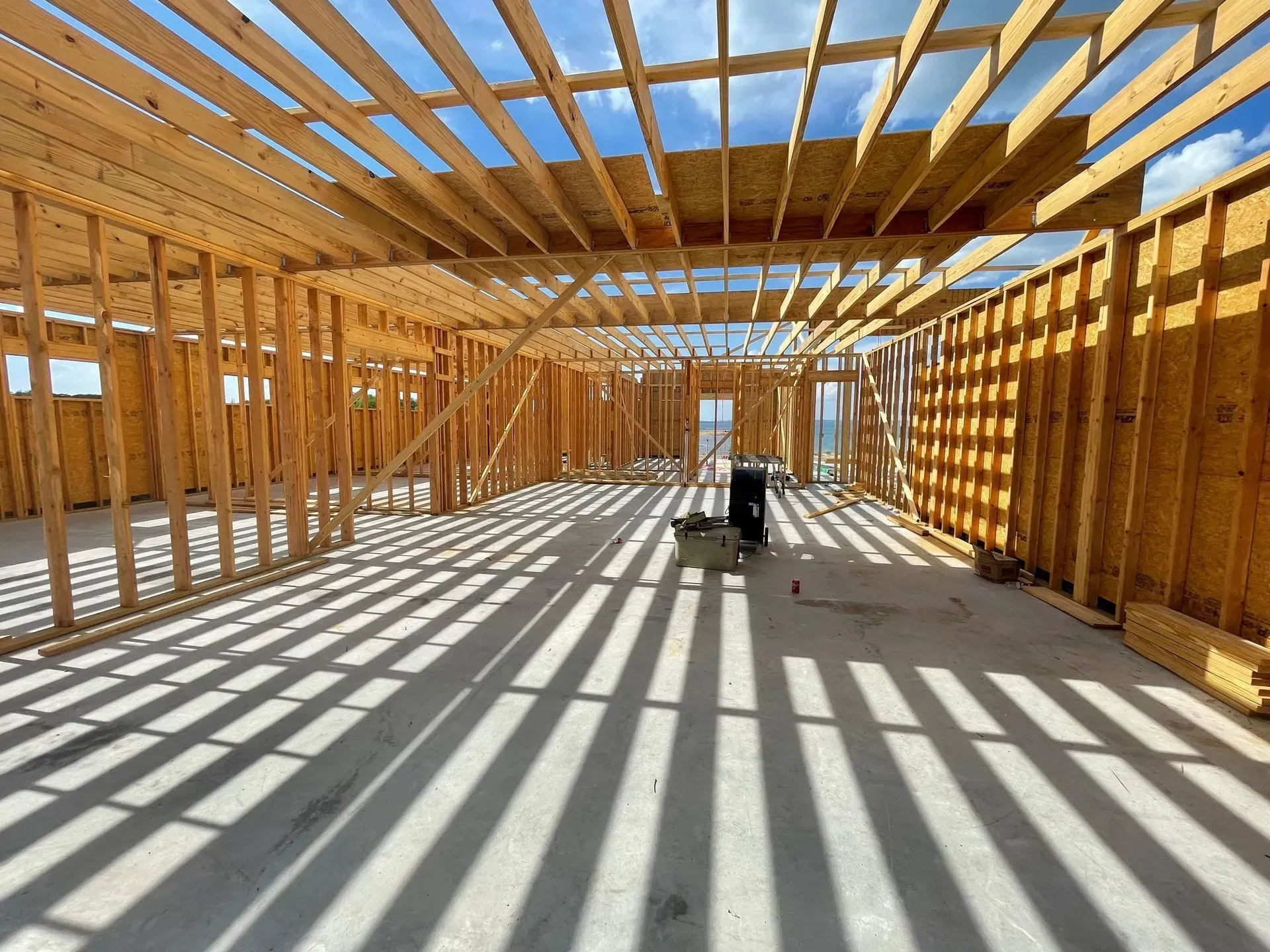 Interior view of a building's wooden framework under construction, with long shadows on the concrete floor.