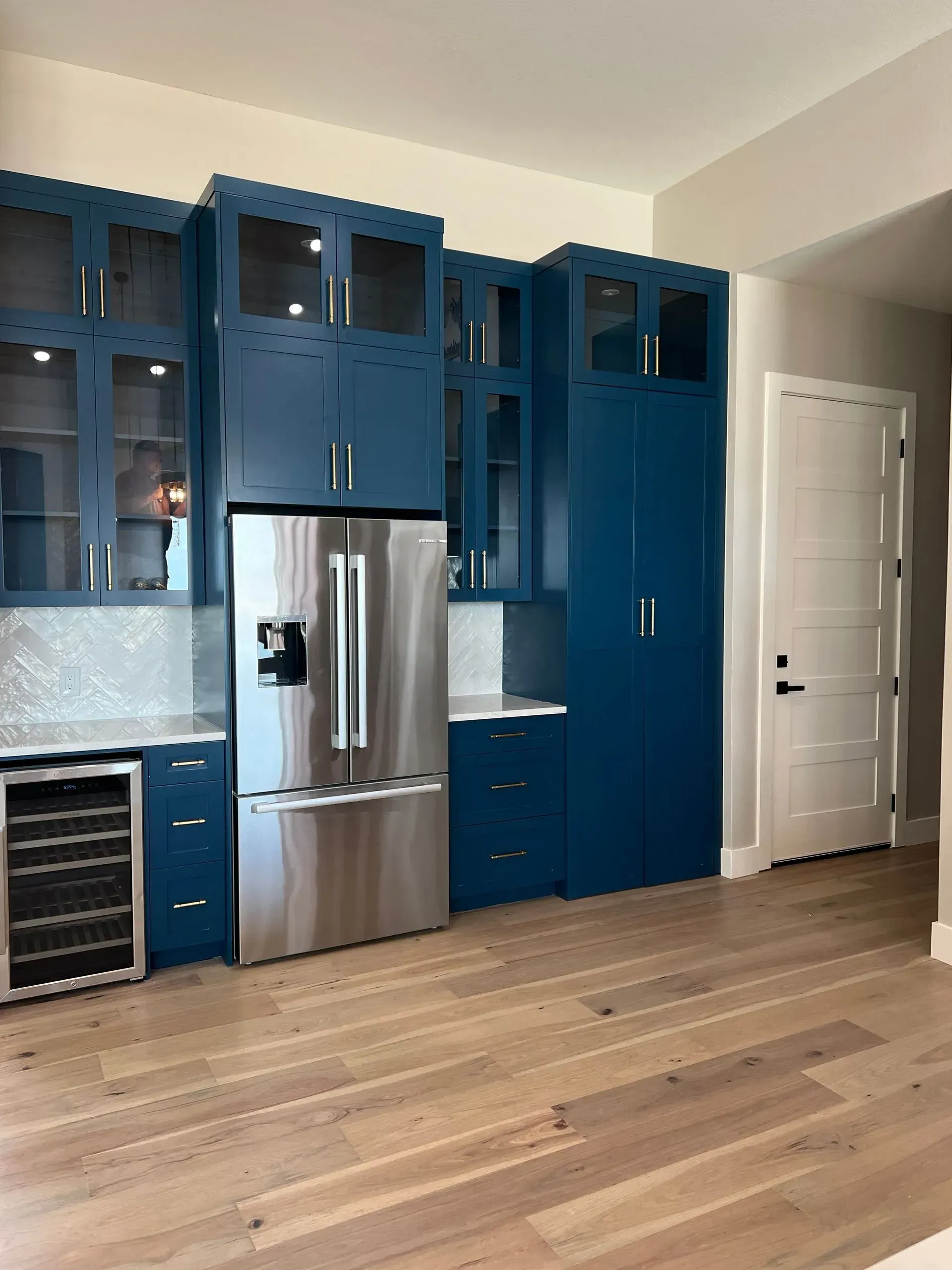 Blue kitchen cabinets surround a stainless steel fridge and wine cooler, set against a light wood floor and white walls.