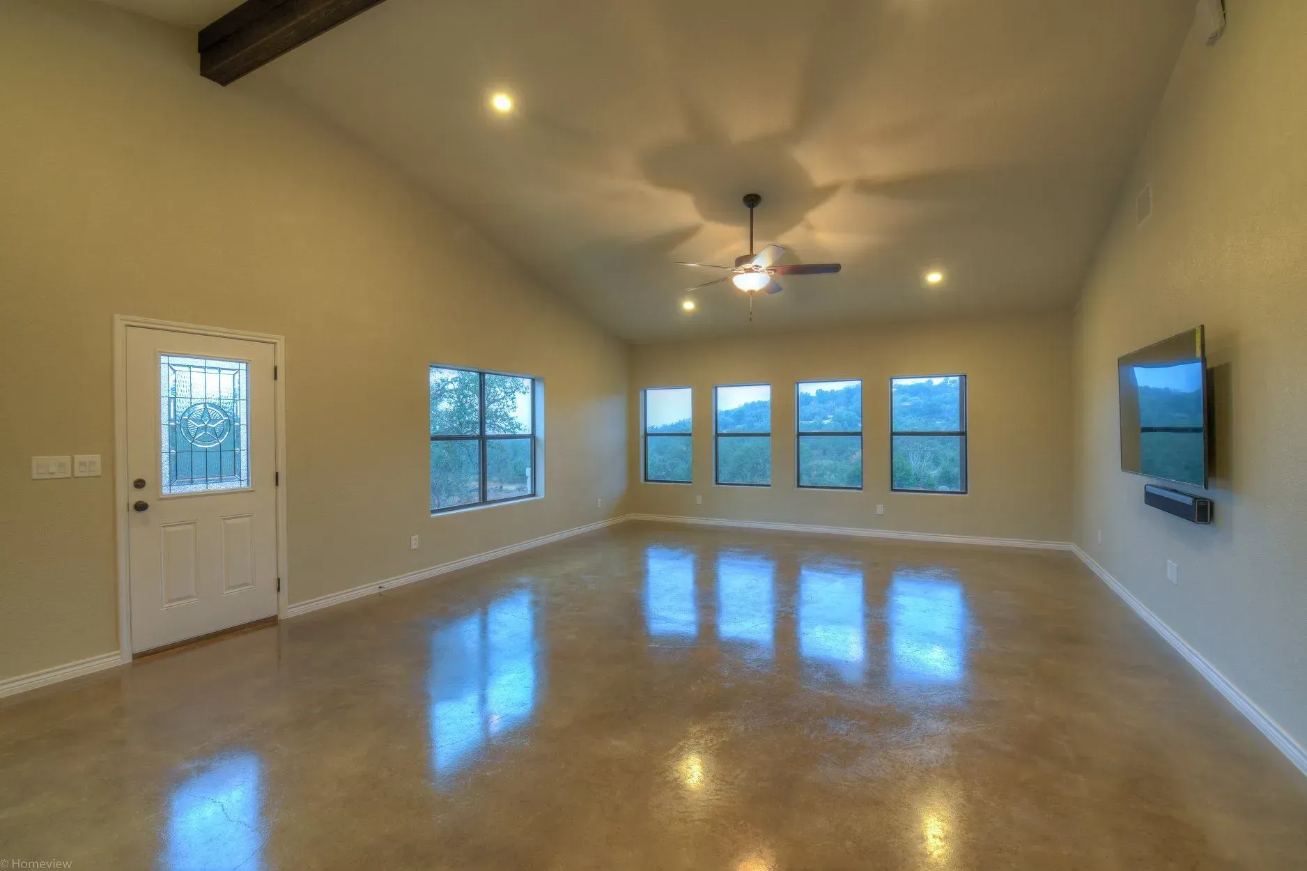 Empty living room with concrete floor, large windows, and mounted TV. Beige walls, natural light.